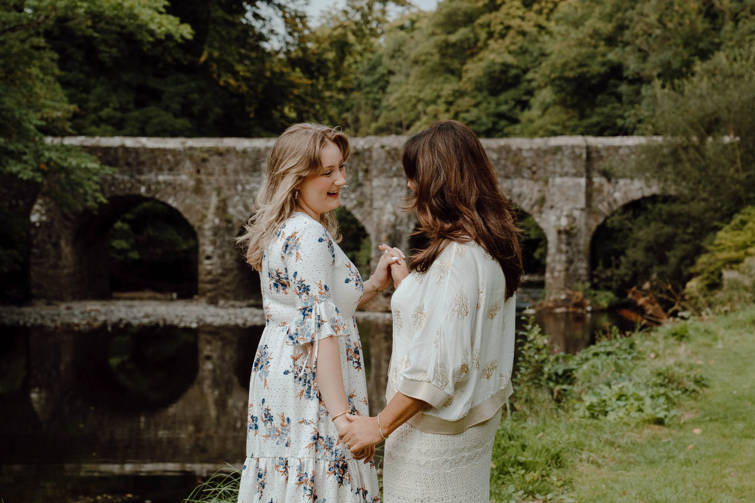 Two women holding hands and smiling by a river with a stone bridge in the background.