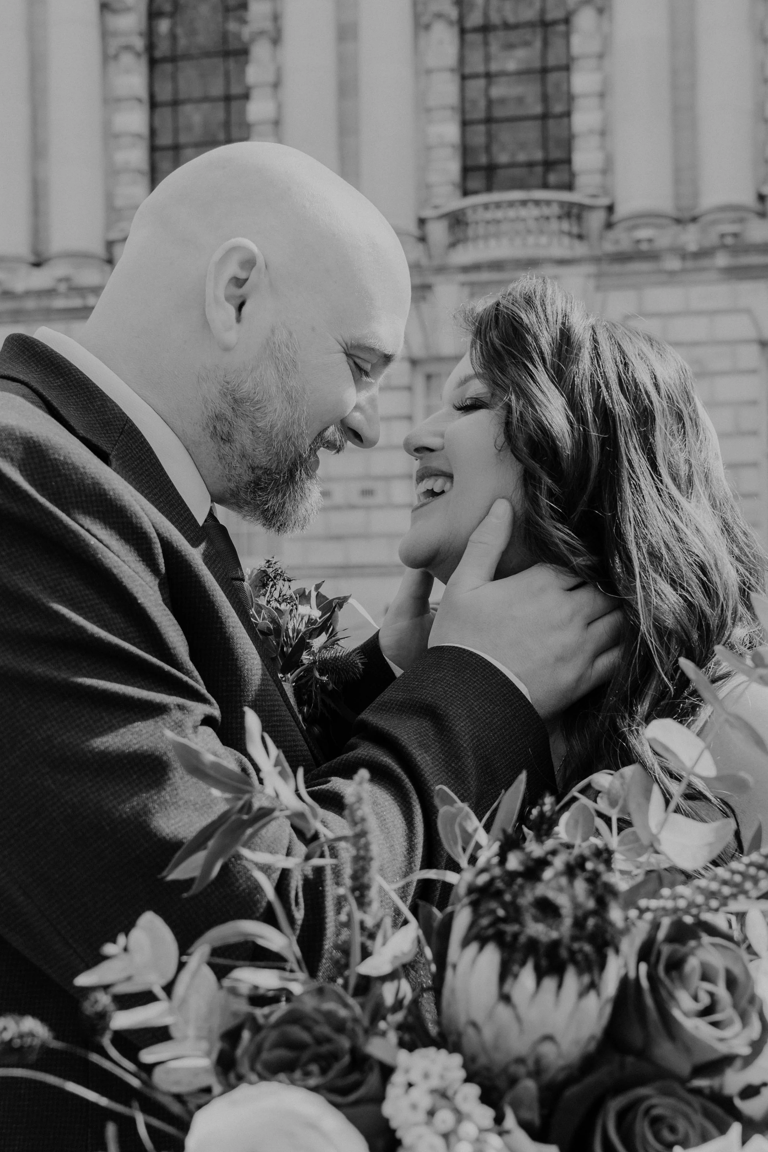 A black and white photo of a couple with their foreheads touching, surrounded by flowers. The man is bald with a beard, and the woman has long, wavy hair. They are smiling and looking at each other affectionately.
