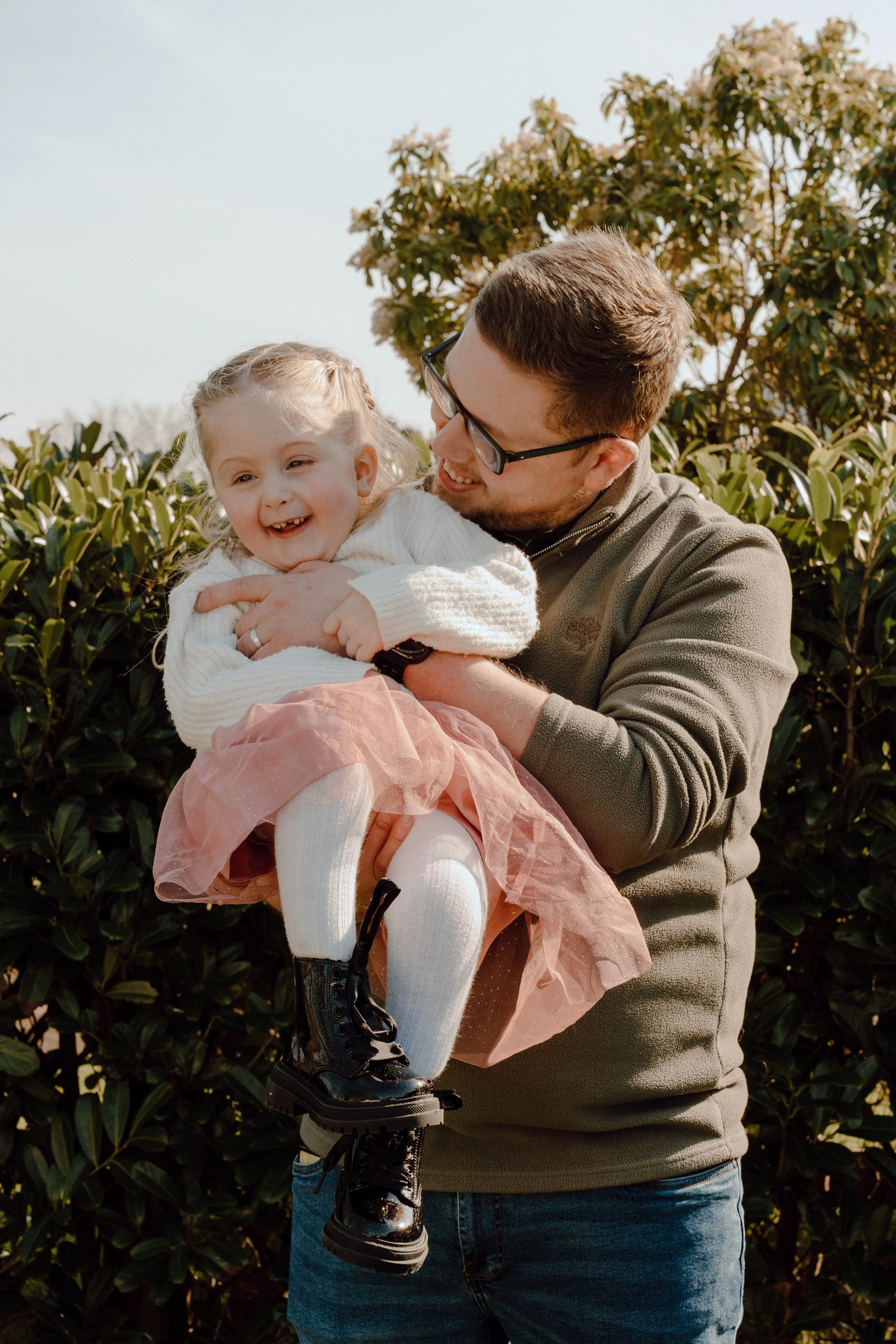 A man holding a smiling young girl outdoors among bushes on a sunny day.