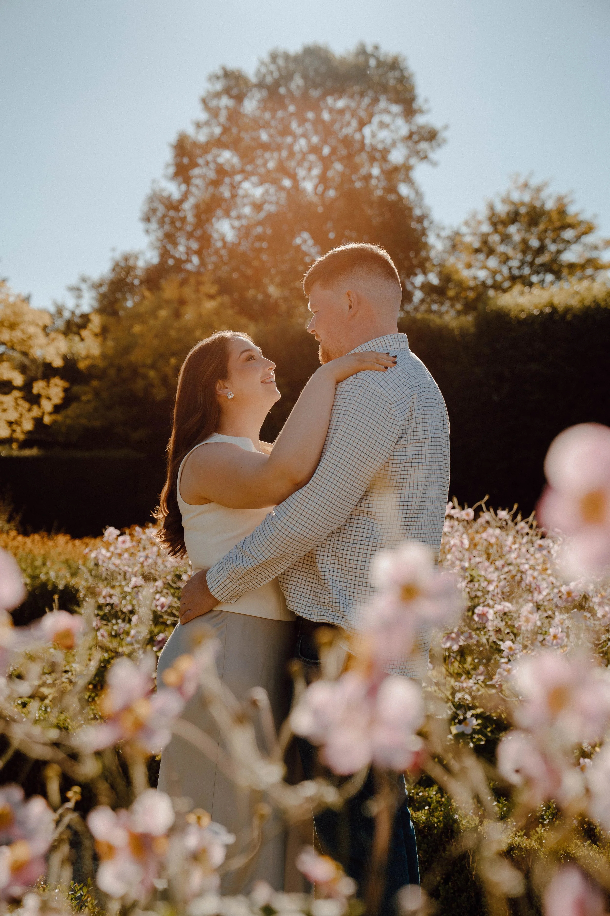 A couple stands in a field of pink flowers, gazing into each other's eyes on a sunny day with trees and a clear sky in the background.