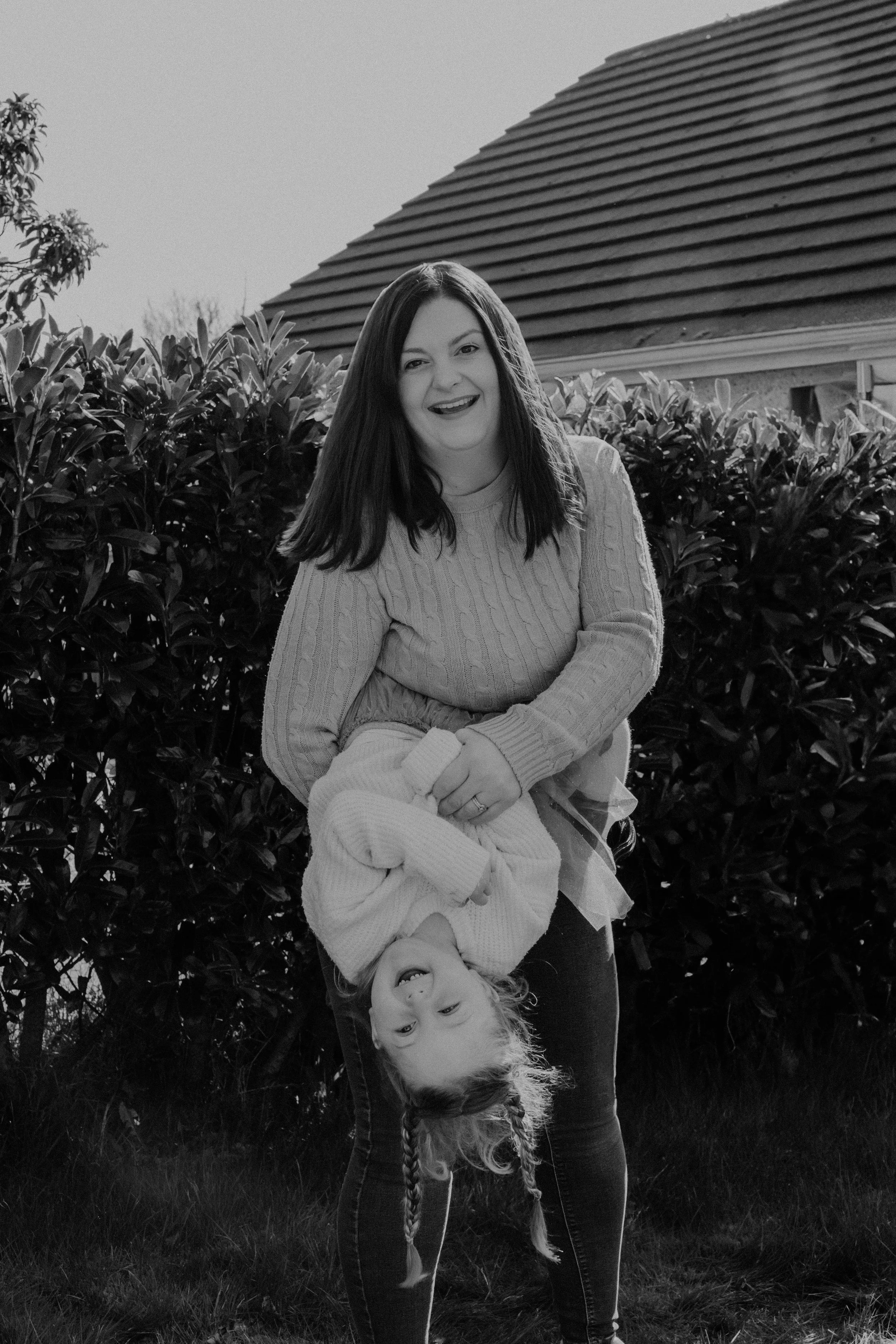 A woman holding a young girl upside down outdoors in front of bushes, smiling at the camera in black and white.