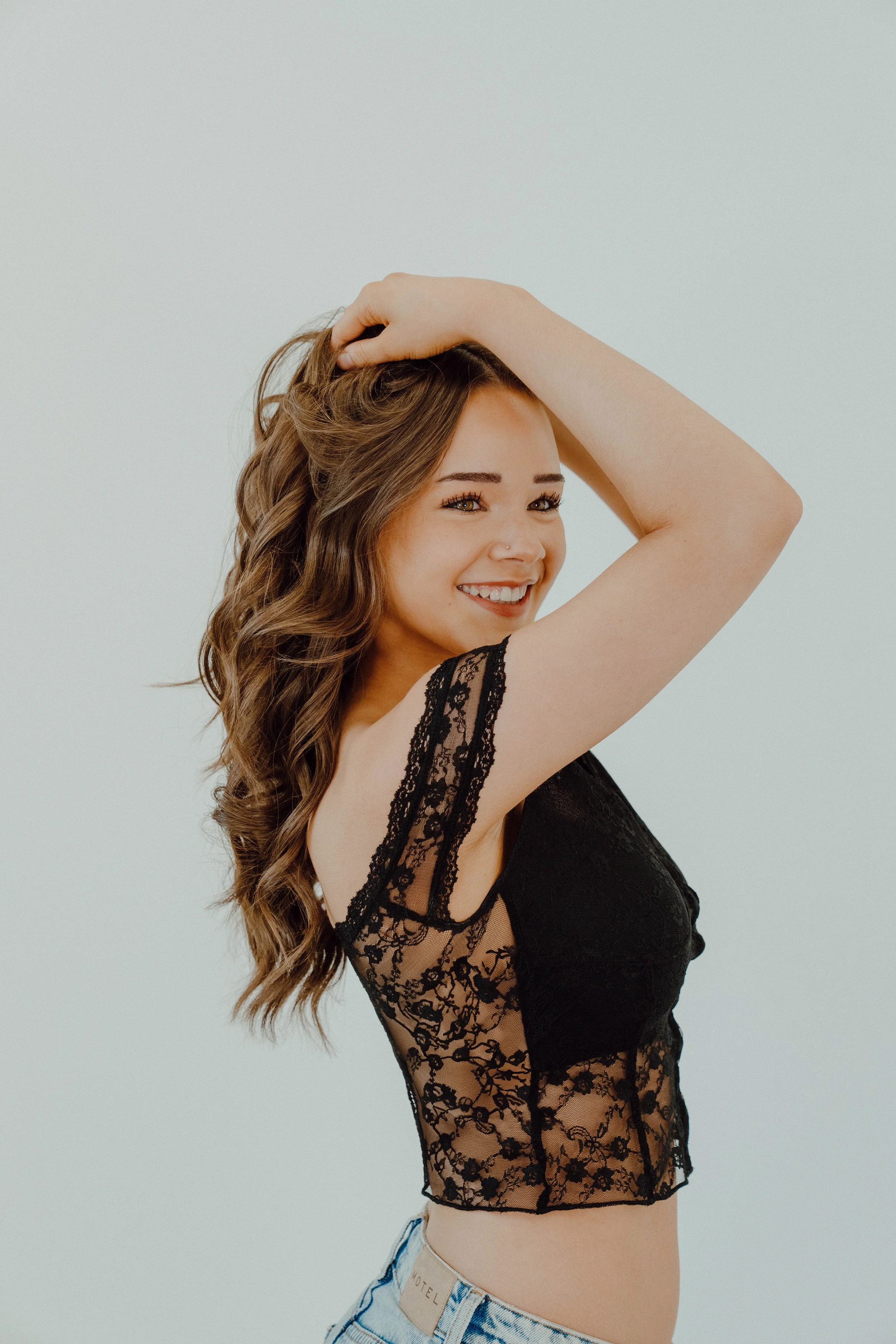 Smiling young woman with long wavy brown hair, wearing a black lace sleeveless top and light denim shorts, posing against a plain white background.