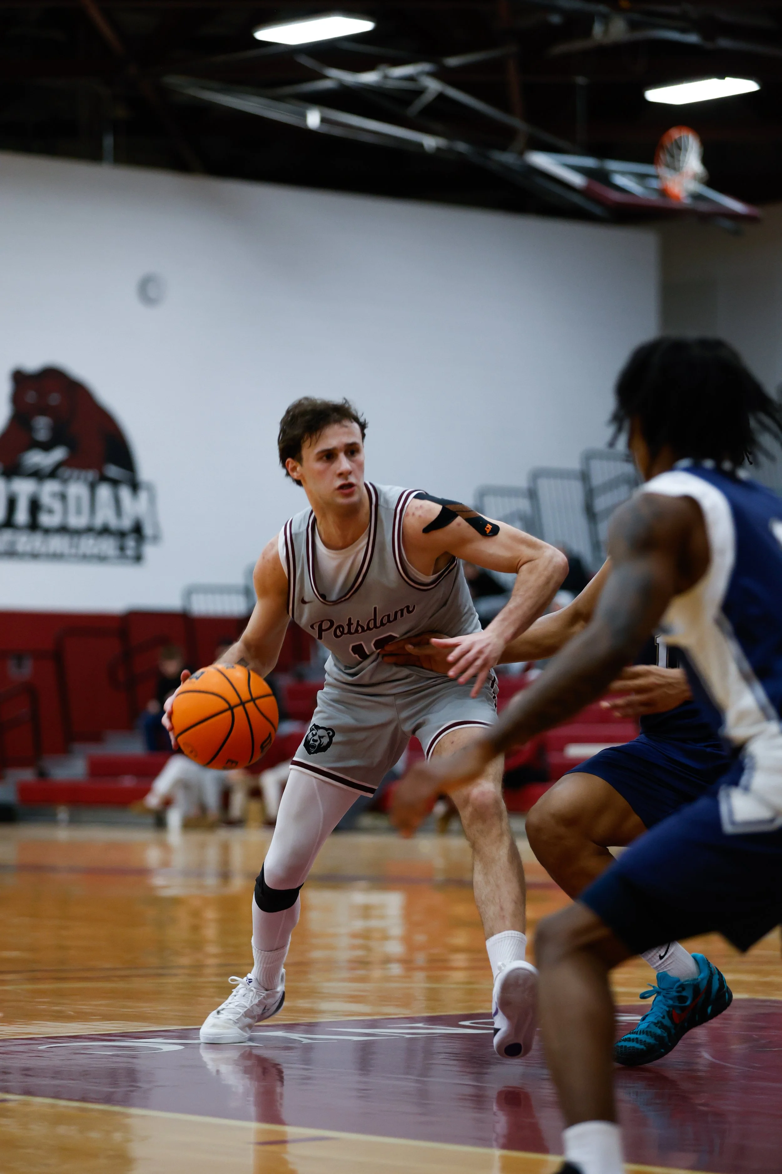 Two basketball players competing during a game in a gymnasium. One player in a gray uniform dribbling the ball, and an opponent in a blue and white uniform defending.
