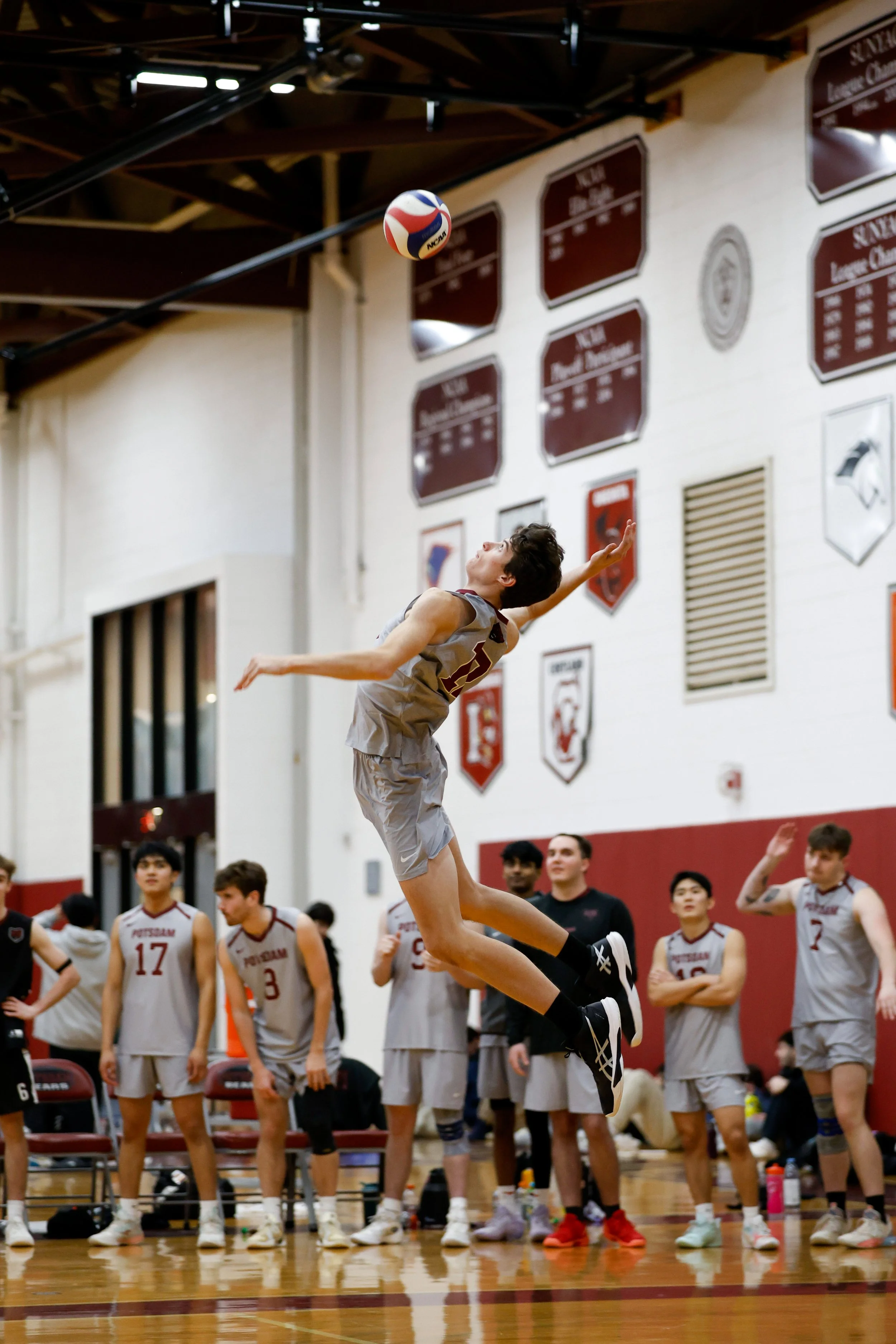 A male basketball player in a grey uniform jumps to serve the volleyball inside a gymnasium, with his teammates watching from behind.