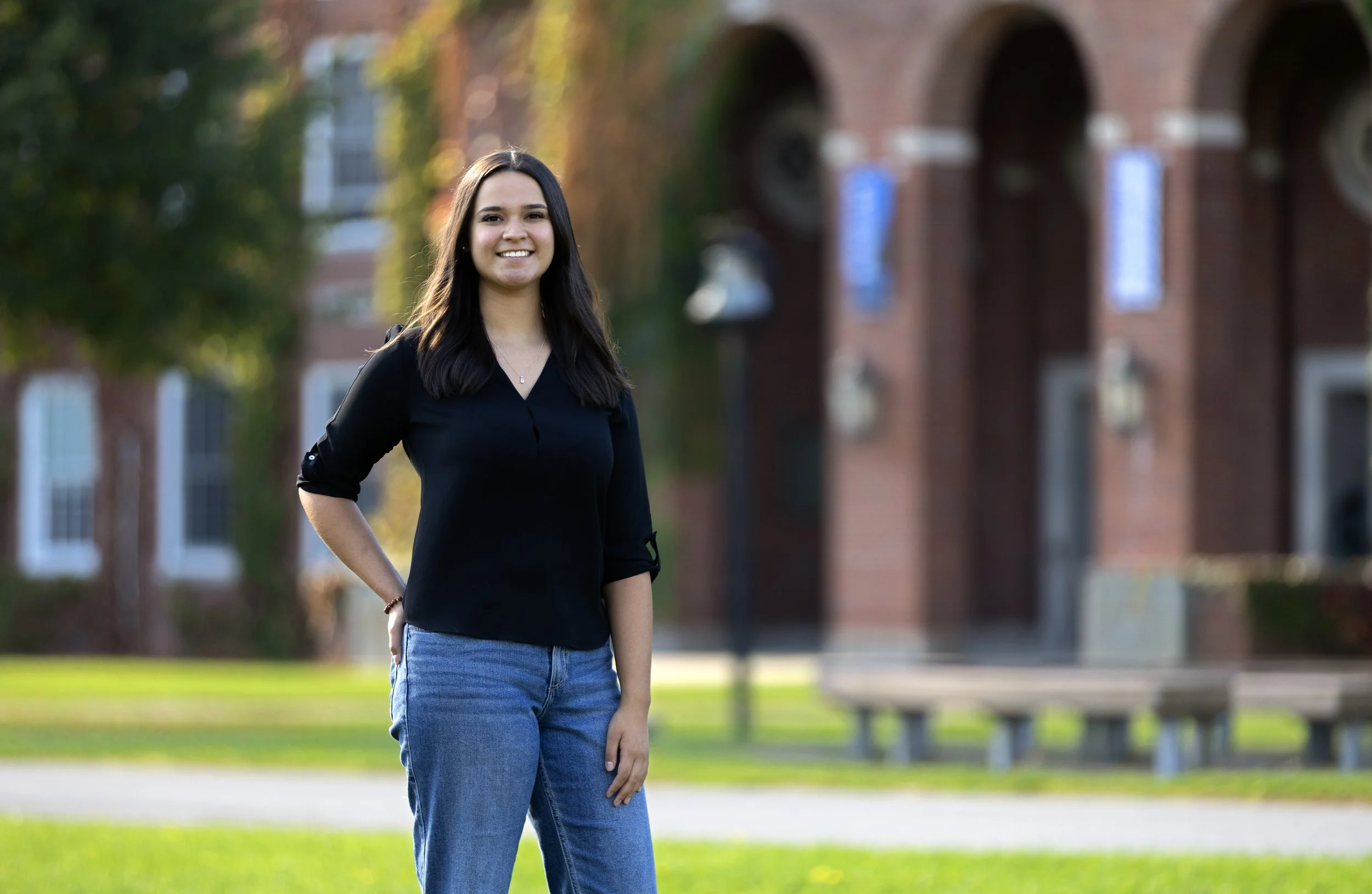 A young woman standing outdoors on a grassy area with a brick building in the background, smiling and looking at the camera.