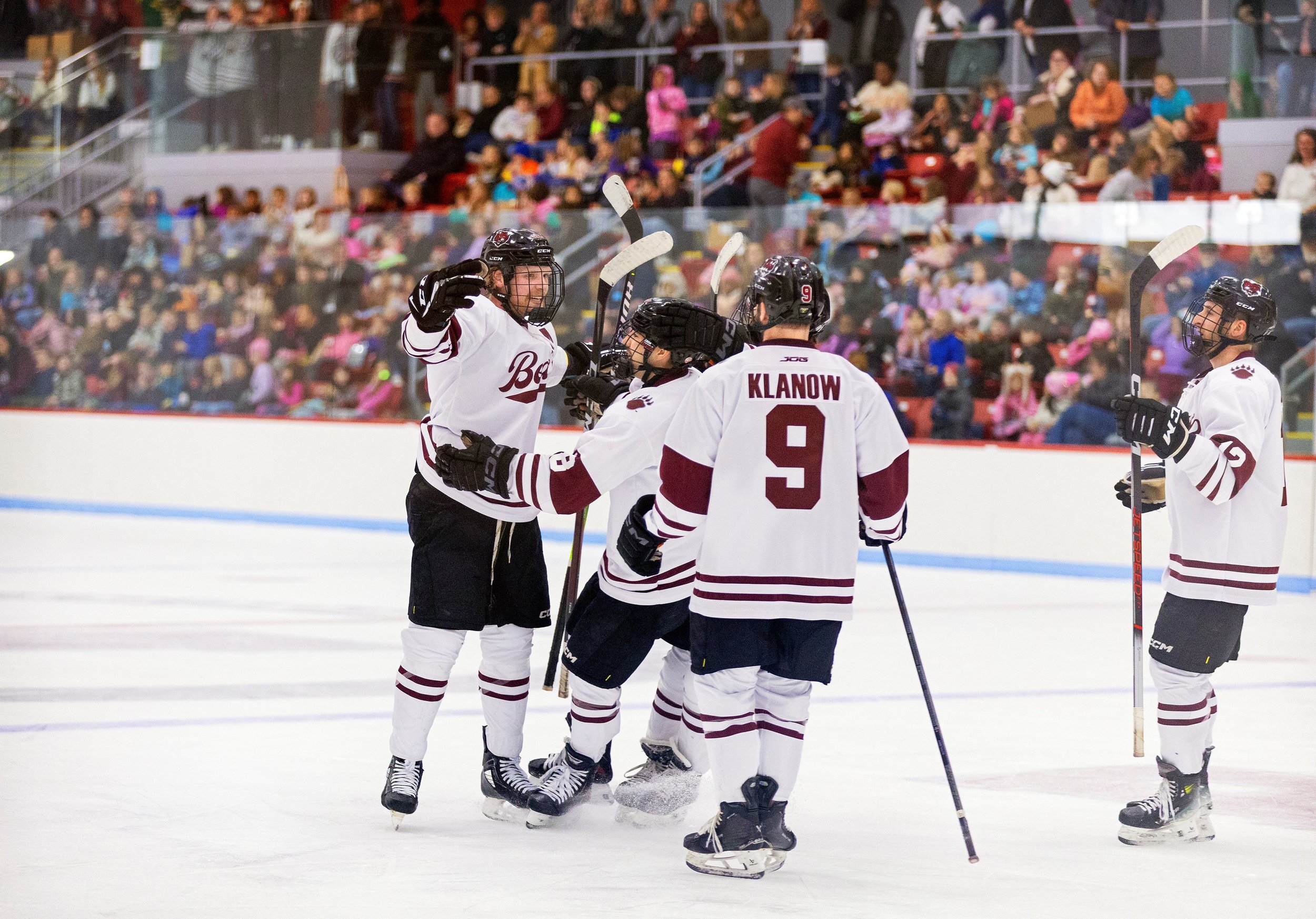 Hockey players in white jerseys celebrating on the ice rink during a game, with a crowd of spectators in the background.