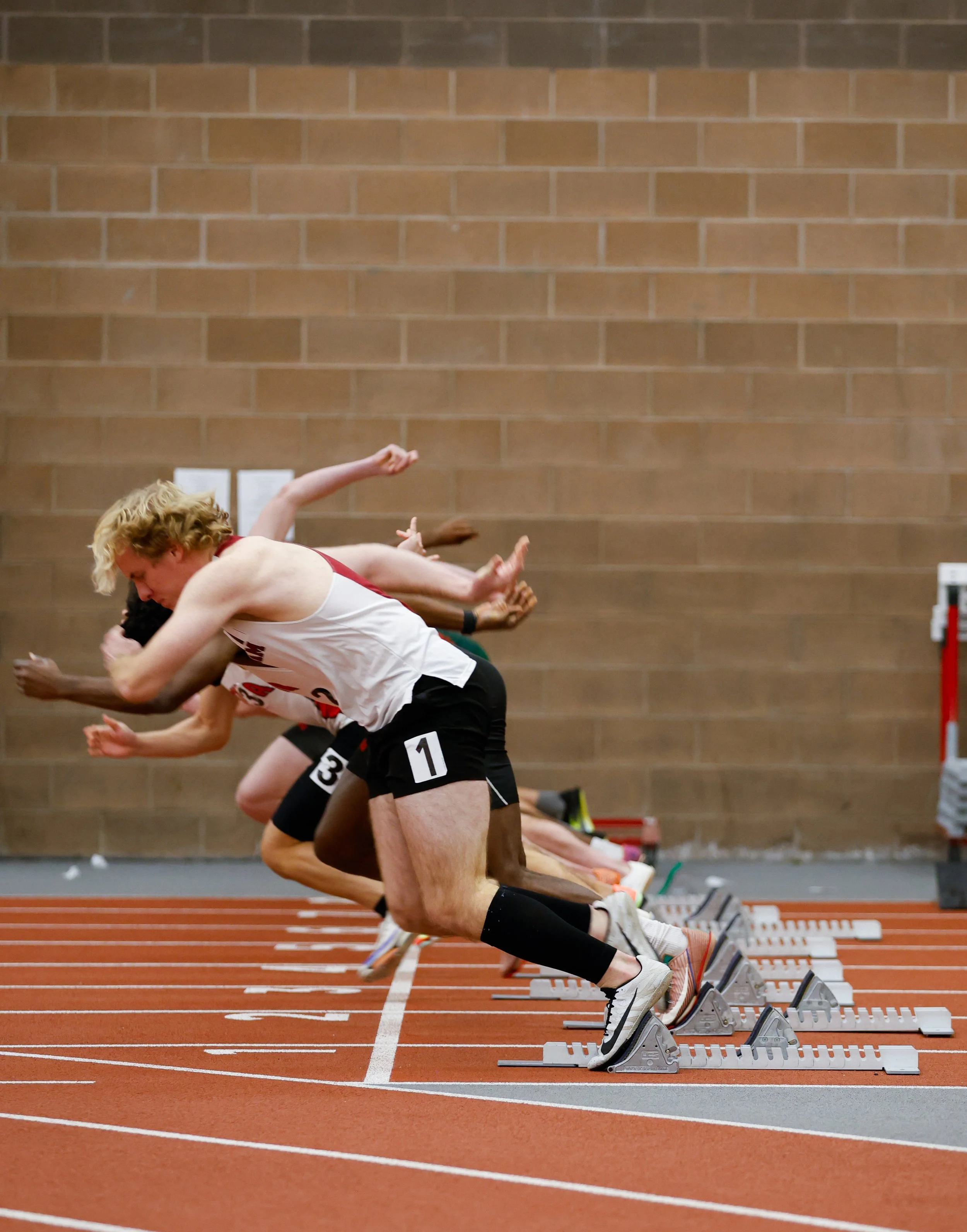 Men starting a race on an indoor track, wearing athletic clothing and race bibs, with hands on the ground at the starting blocks.