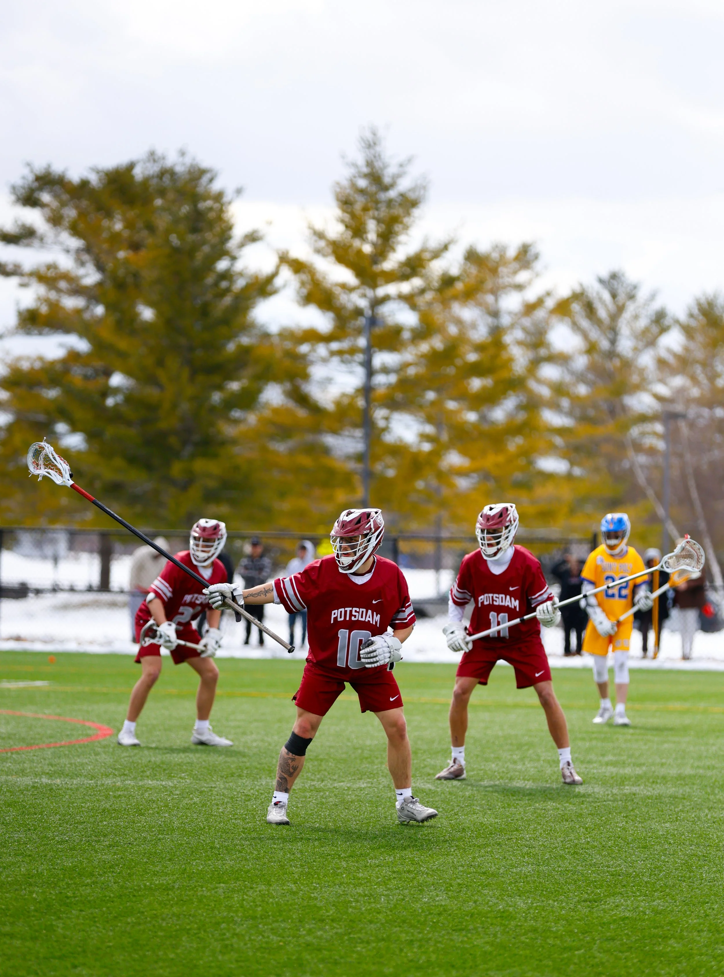 Lacrosse players in a game, wearing helmets and uniforms, with two players in red and one in yellow, on a grassy field with trees in the background.