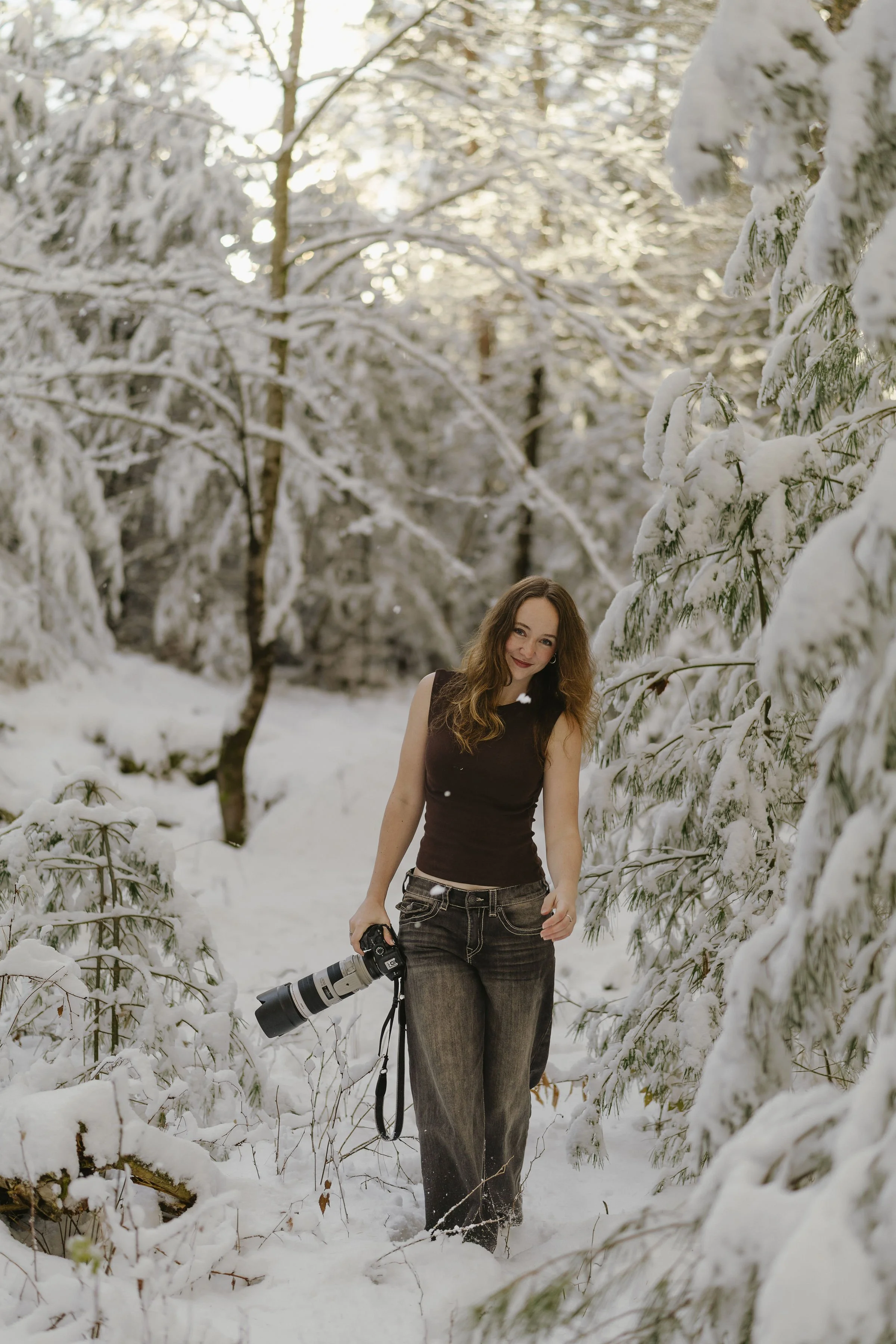 A woman walking through a snow-covered forest with a camera hanging from her neck.