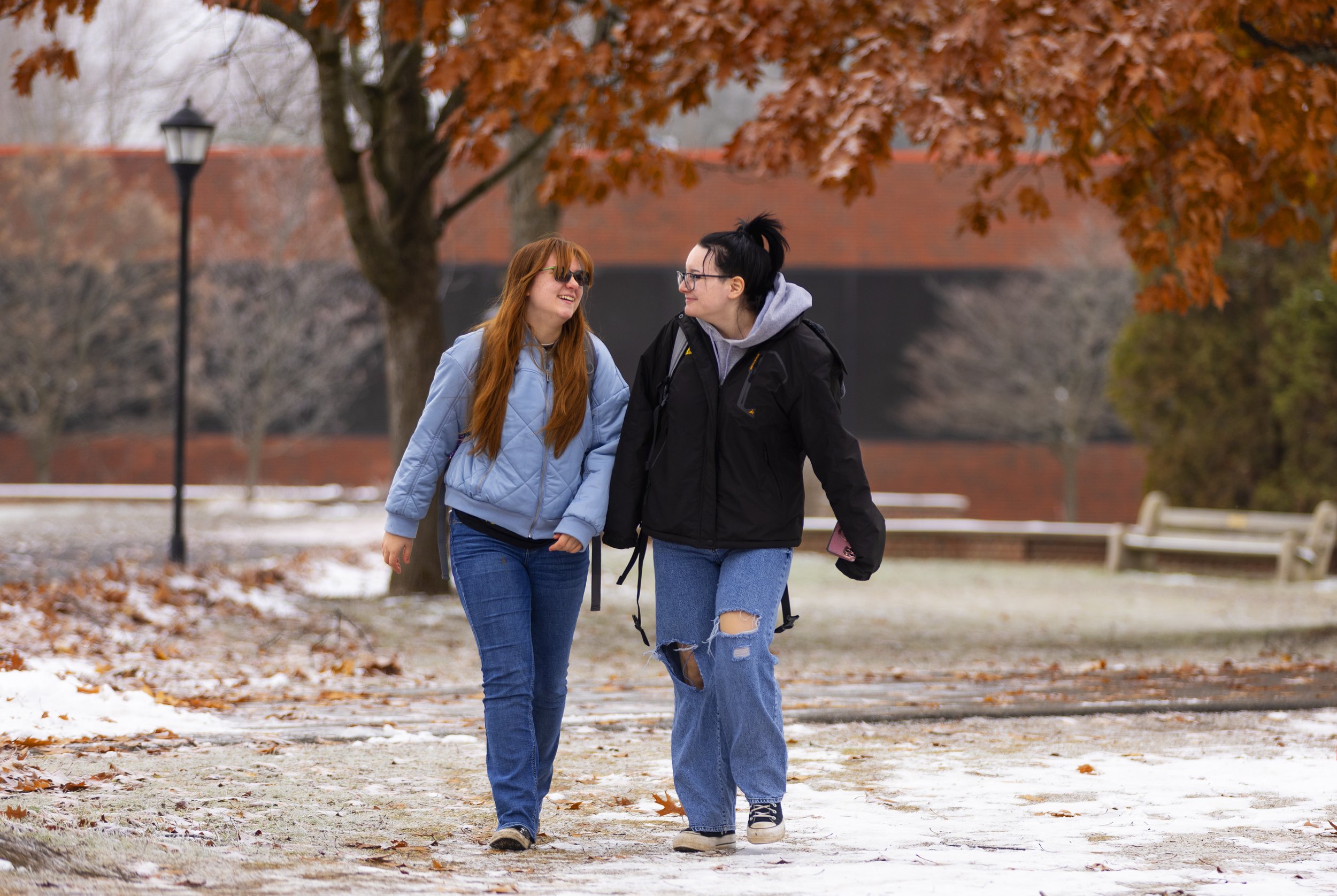 Two women walking and talking outdoors in a park during late fall or early winter, with trees and benches in the background, some snow on the ground.