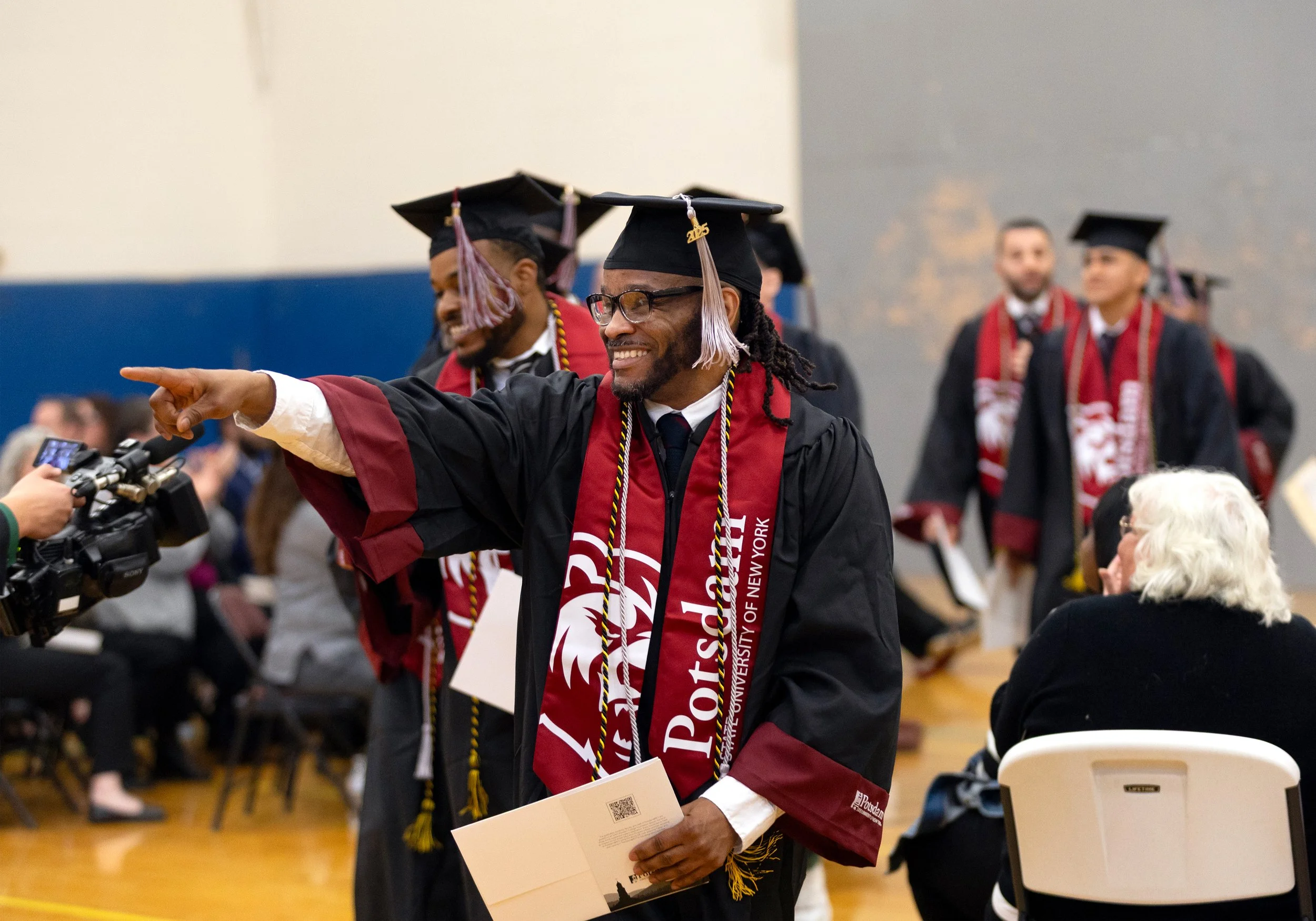 Graduates in caps and gowns at a commencement ceremony, one smiling man with glasses and dreadlocks is pointing and holding a program, others behind are clapping and smiling, indoor setting with audience seated.