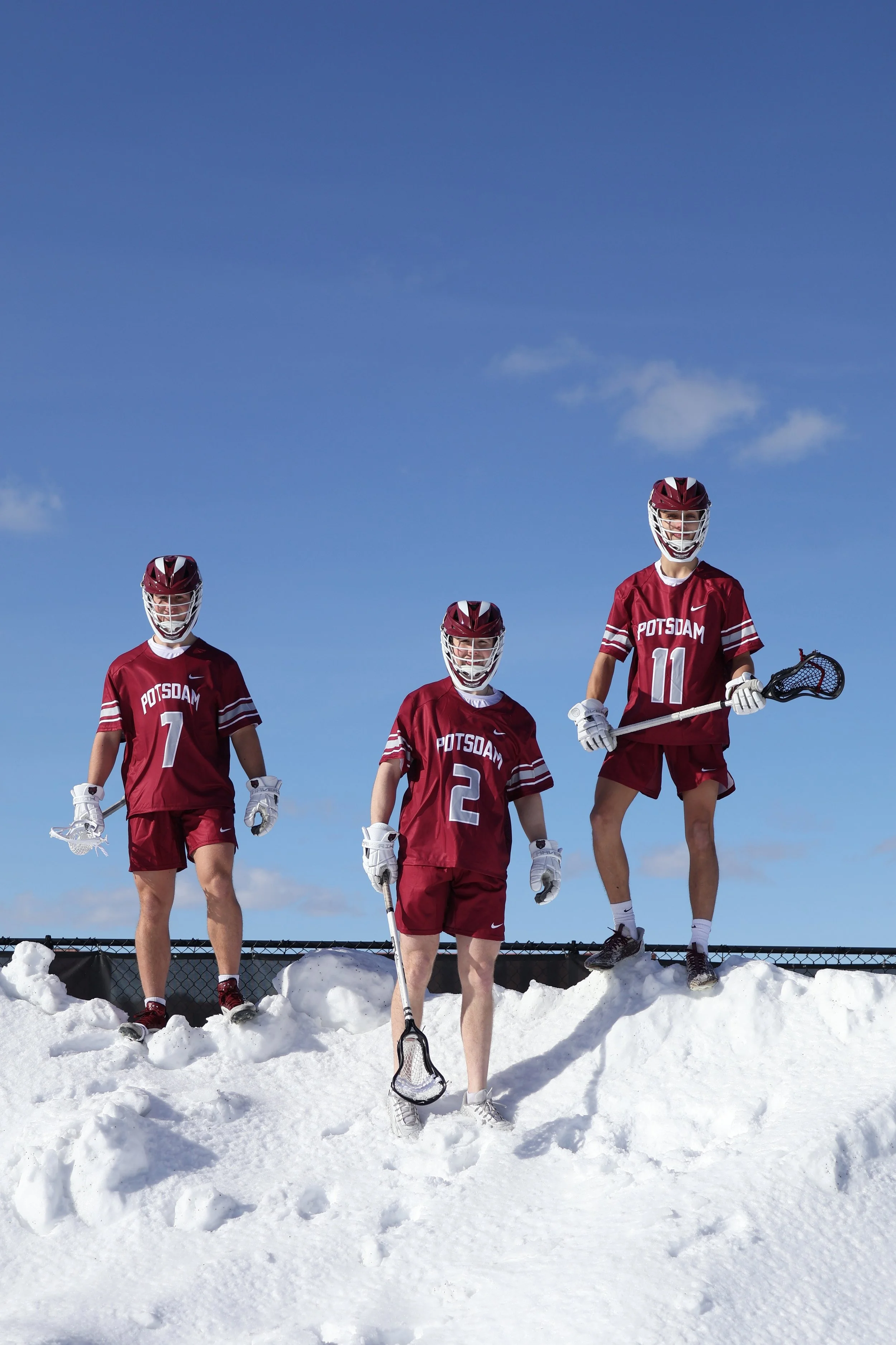 Three lacrosse players in maroon uniforms with 'POTSDAM' on them, standing on snow against a blue sky. One holds a lacrosse stick, another is holding a lacrosse stick with a net, and the third has a lacrosse stick with a net, all wearing helmets and 