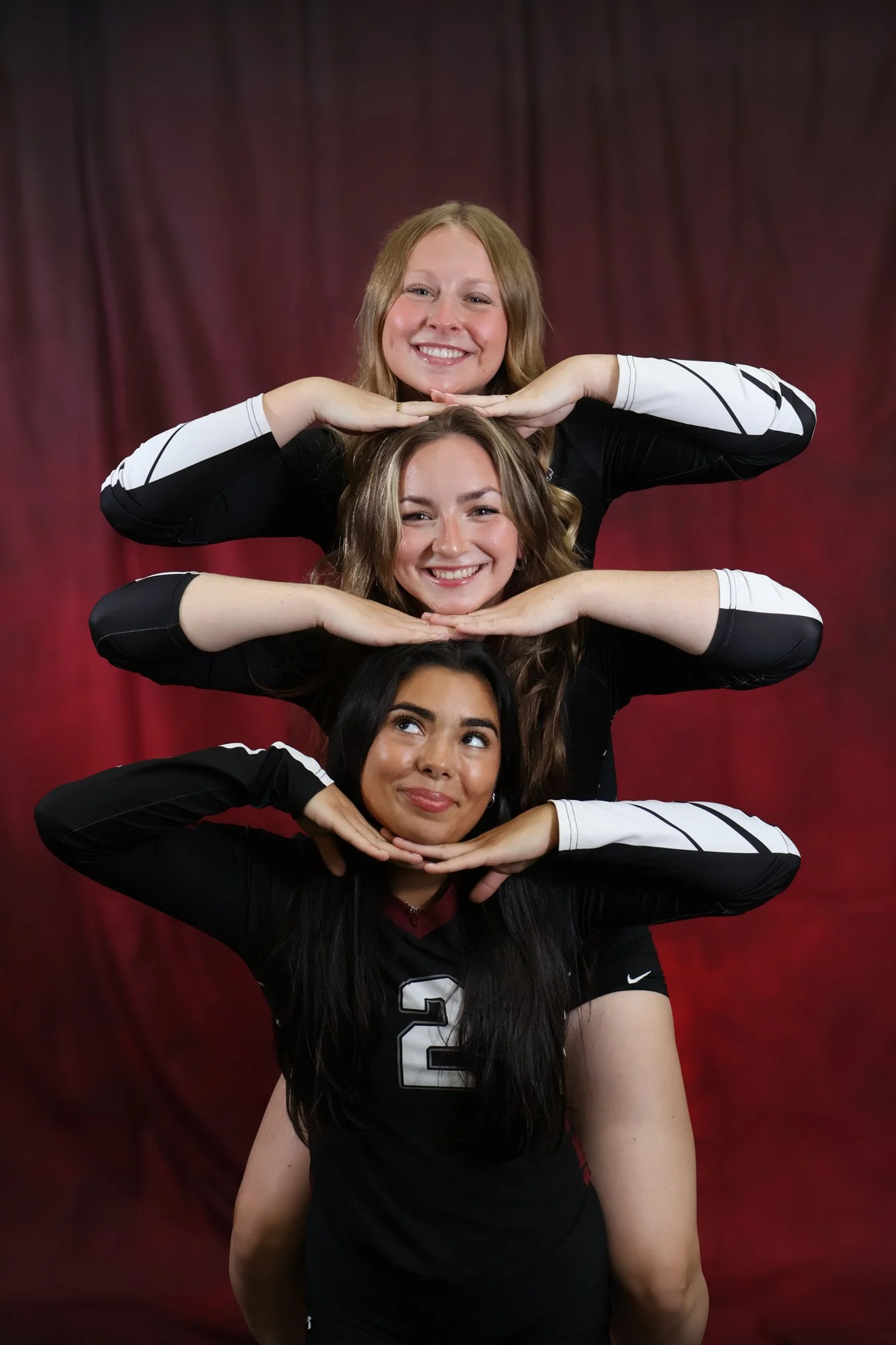 Three women in athletic uniforms stacked on top of each other, smiling and posing with their hands under their chins against a red curtain background.
