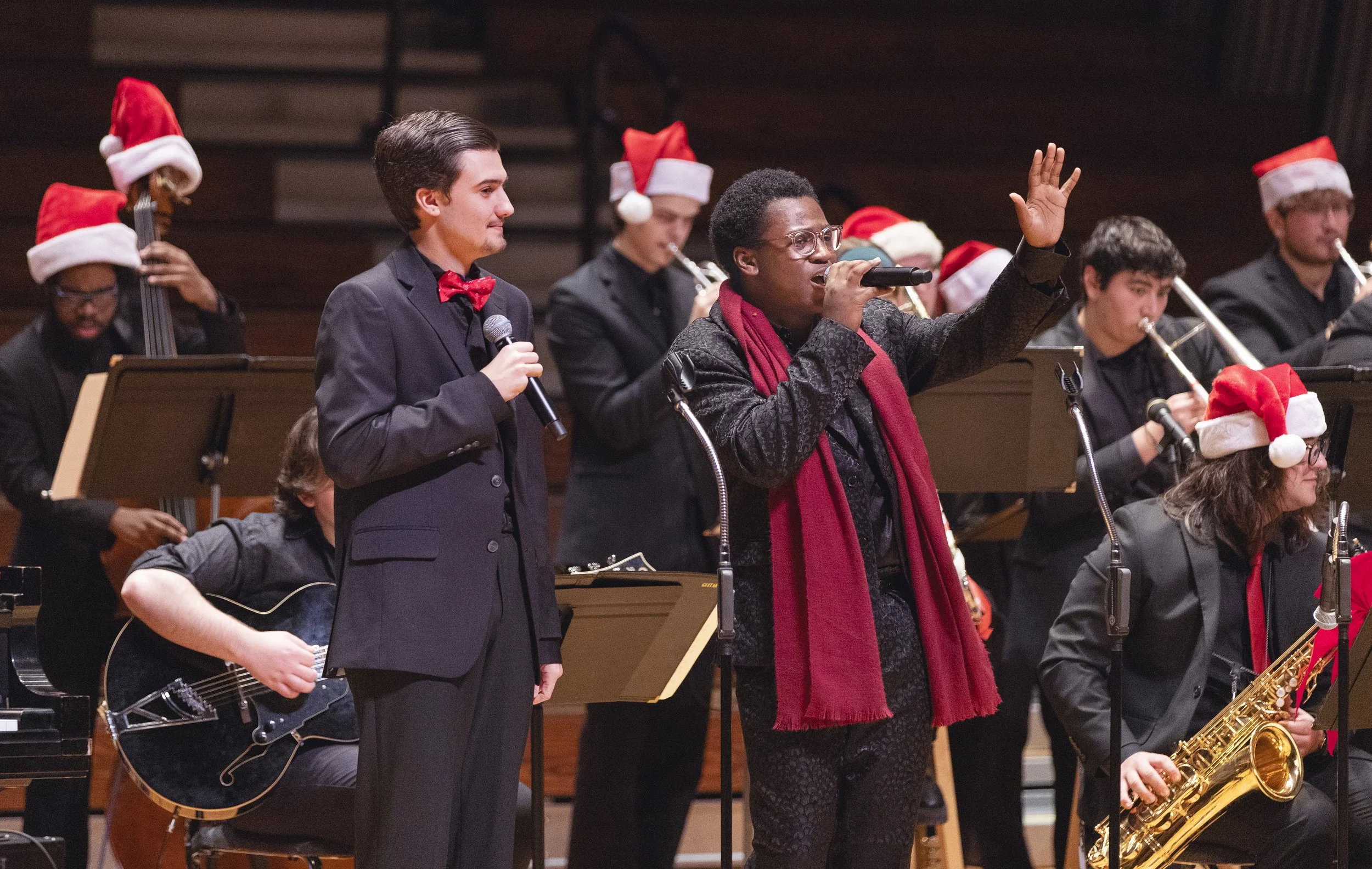 Music performers in Christmas hats, with one person singing into a microphone and another person holding a microphone, surrounded by musicians playing instruments, on a stage during a holiday concert.