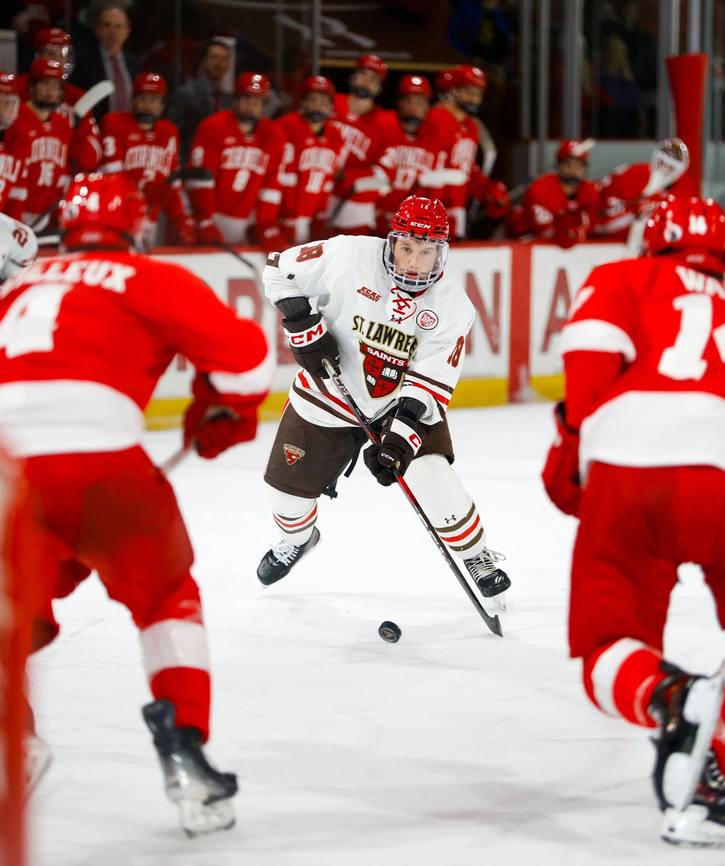 Hockey players in red and white uniforms compete for the puck on the ice, with team members watching from the bench in the background.