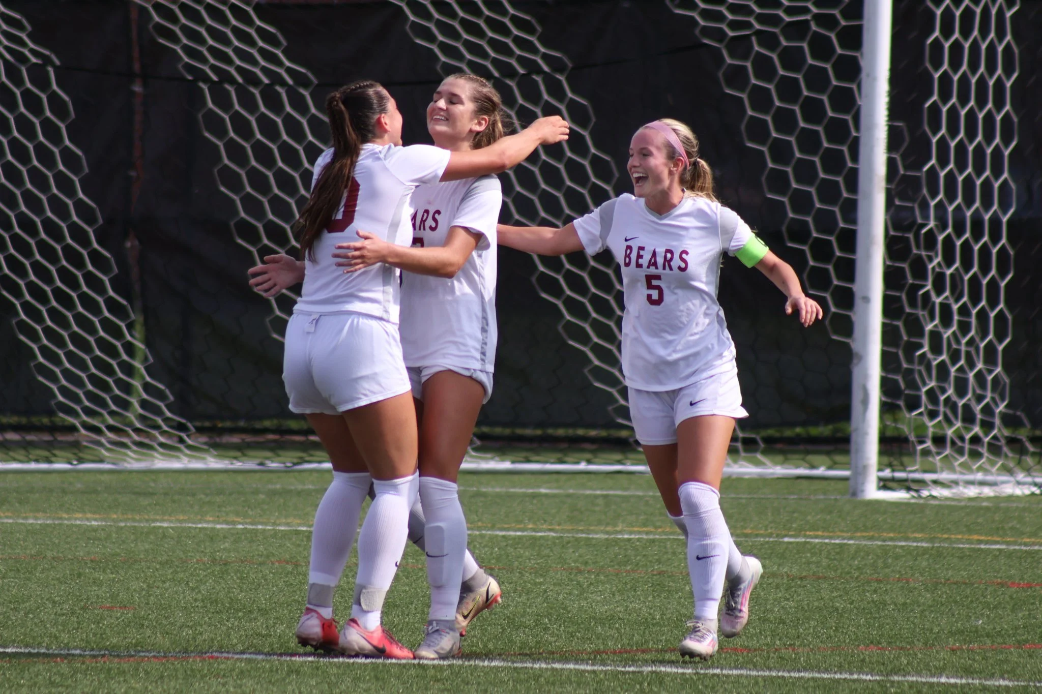 Four female soccer players in white uniforms celebrating on the field, near the goal net, with one wearing a green captain's armband.
