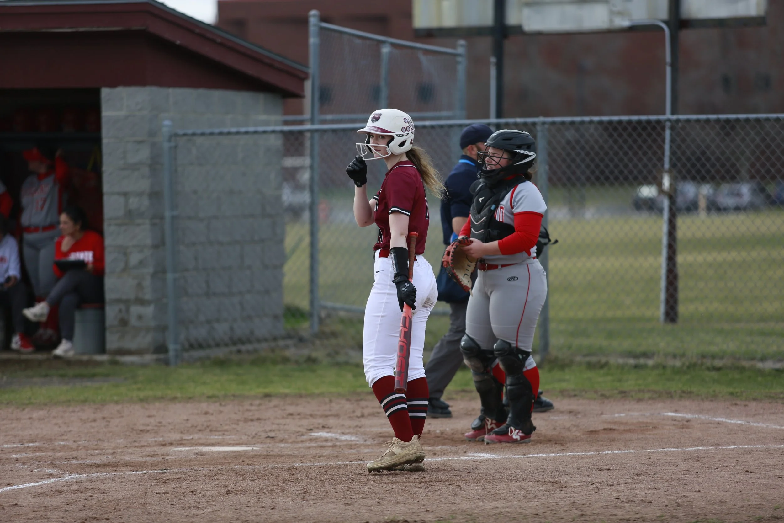 A female softball player in a maroon and white uniform standing at home plate during a game, holding a bat, with a catcher in gray and red gear and a helmet standing behind her. In the background, there are people watching behind a chain-link fence.