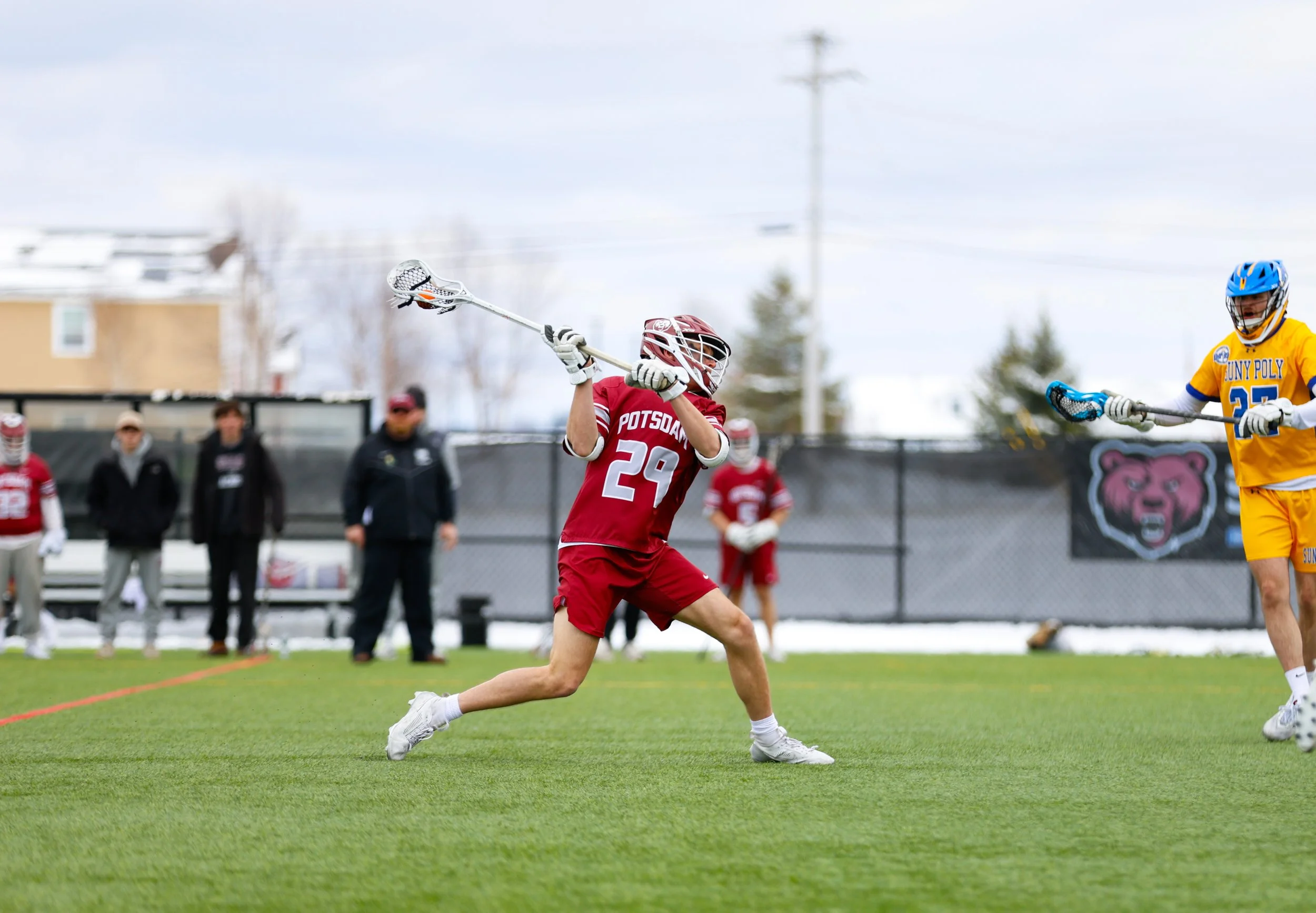 Lacrosse player in red uniform with number 29 tries to catch or block the ball from an opponent in yellow uniform on a green field, with spectators and a black fence in the background.