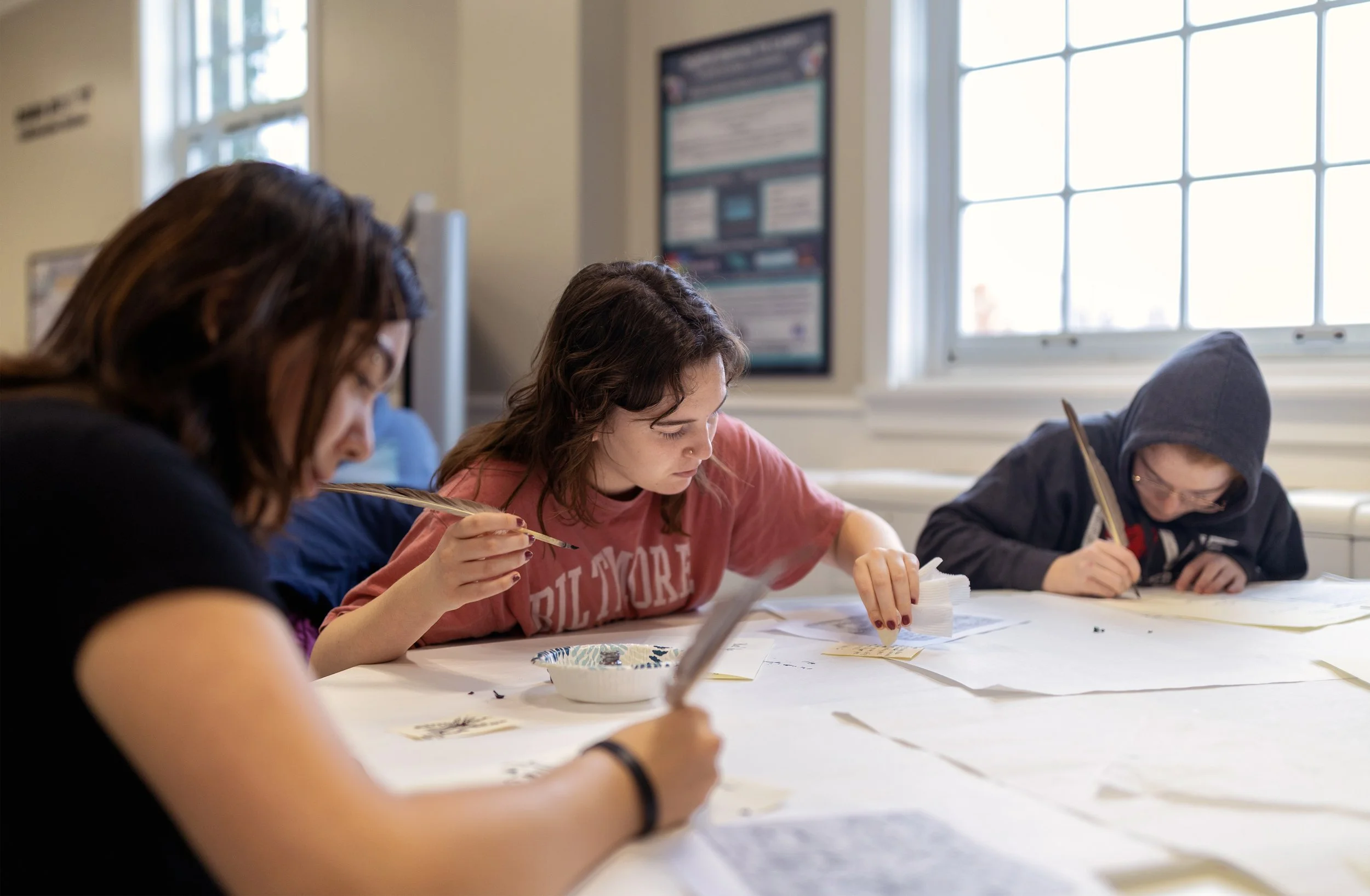 Three children sitting at a table working on art projects with feathers and paper, near a window in a classroom.