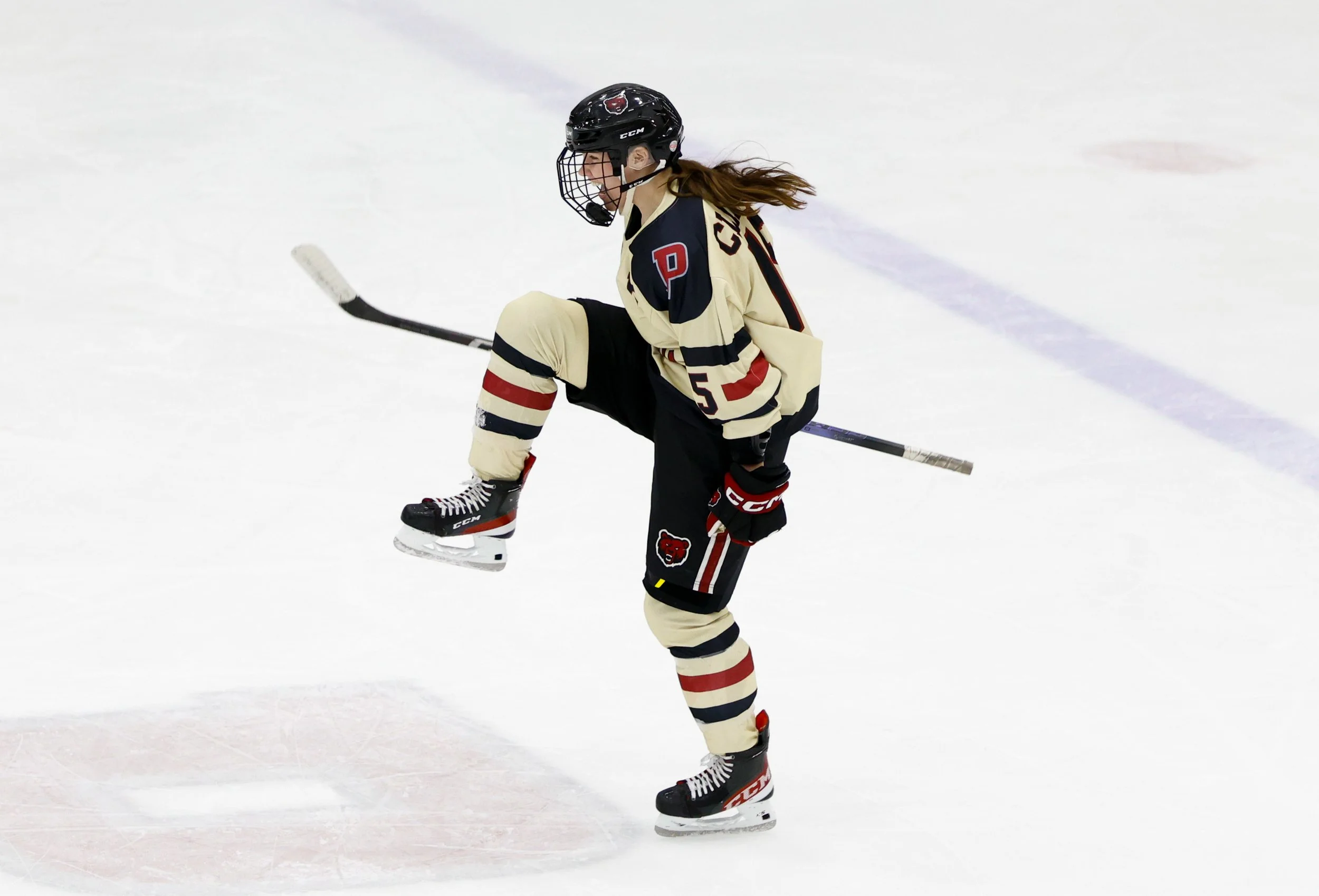 Female hockey player in a beige uniform with black and red accents, wearing a helmet, gloves, and skates, celebrates a goal on the ice rink.