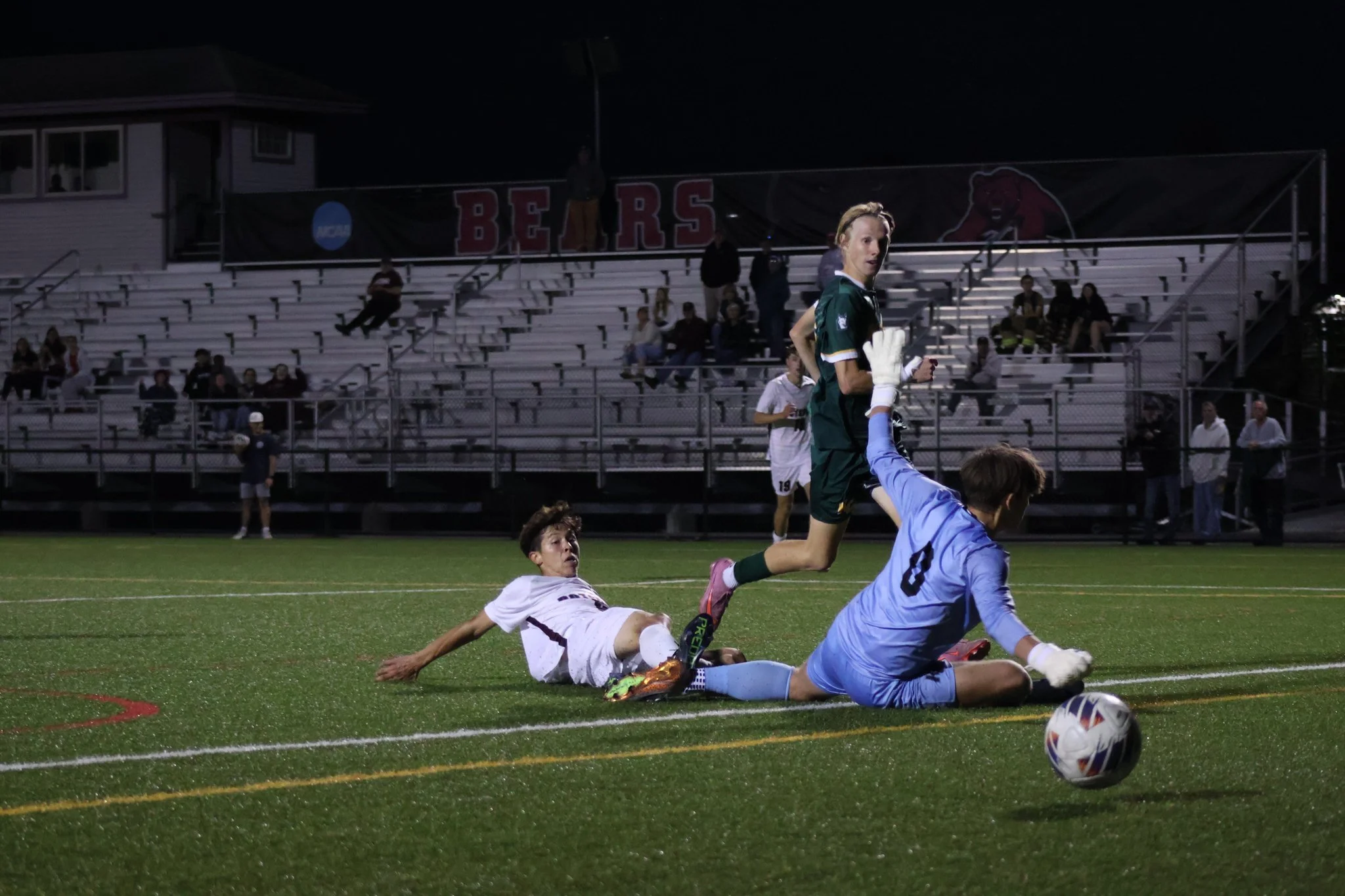 A soccer game at night with a player in green jumping over a falling opponent in white while the goalkeeper in blue dives for the ball.