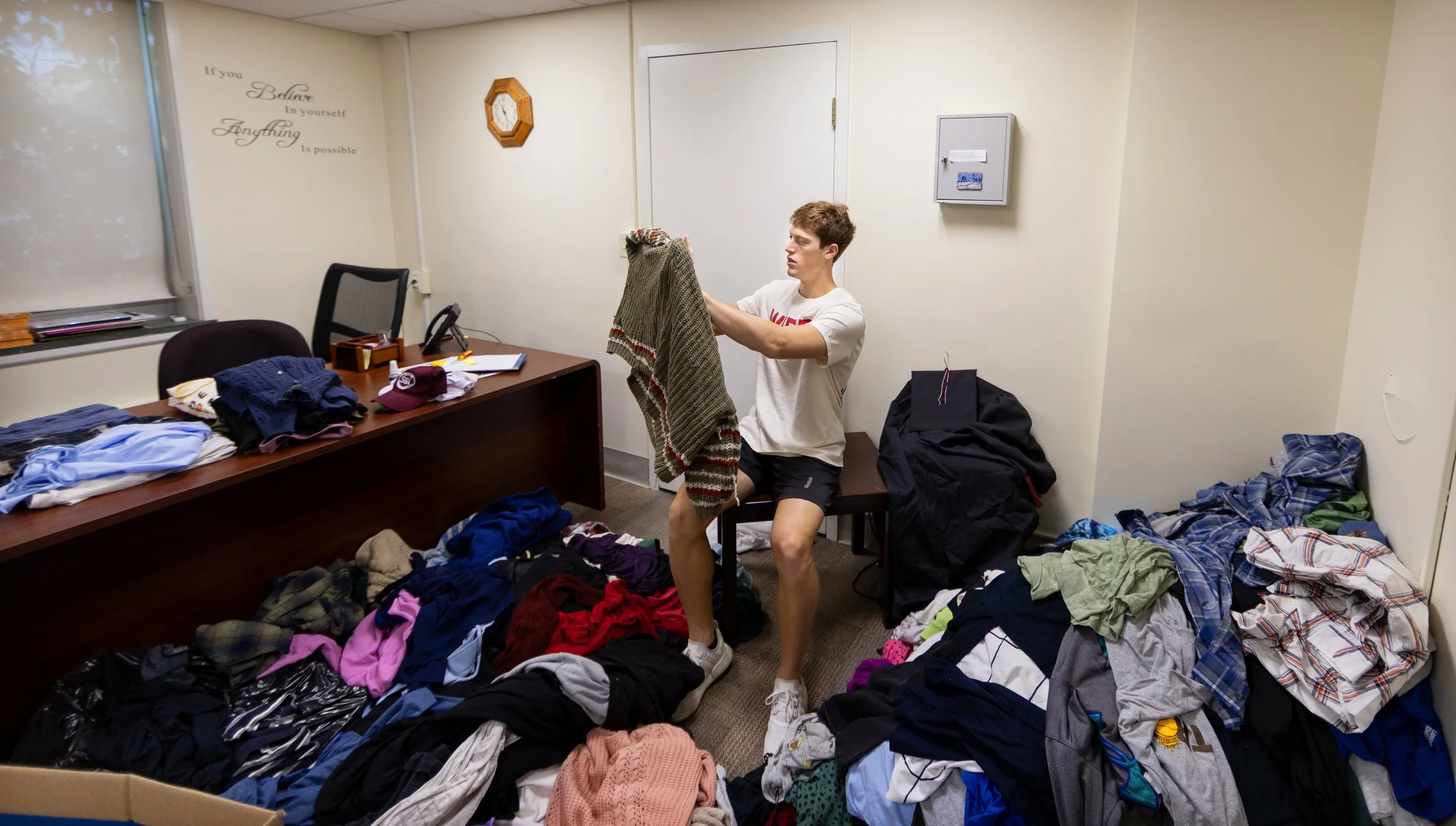 A young man in a white t-shirt and black shorts folding a striped sweater inside a room filled with piles of clothes on the floor and on a desk.