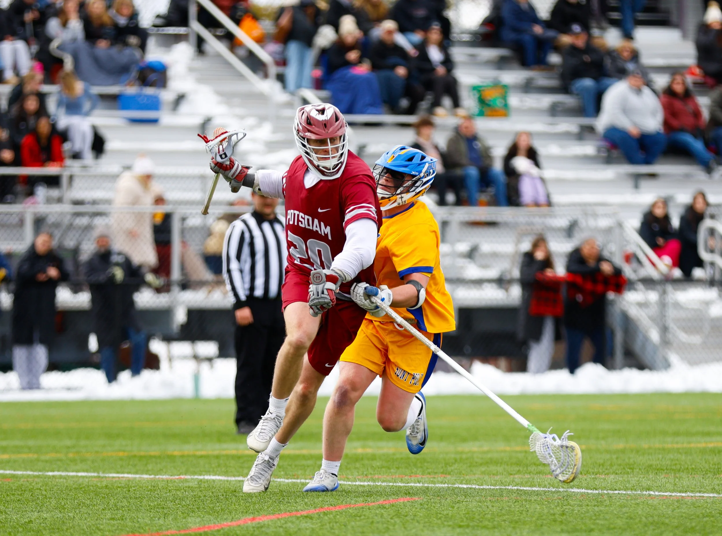 Lacrosse players in action on the field, with spectators on bleachers in the background.