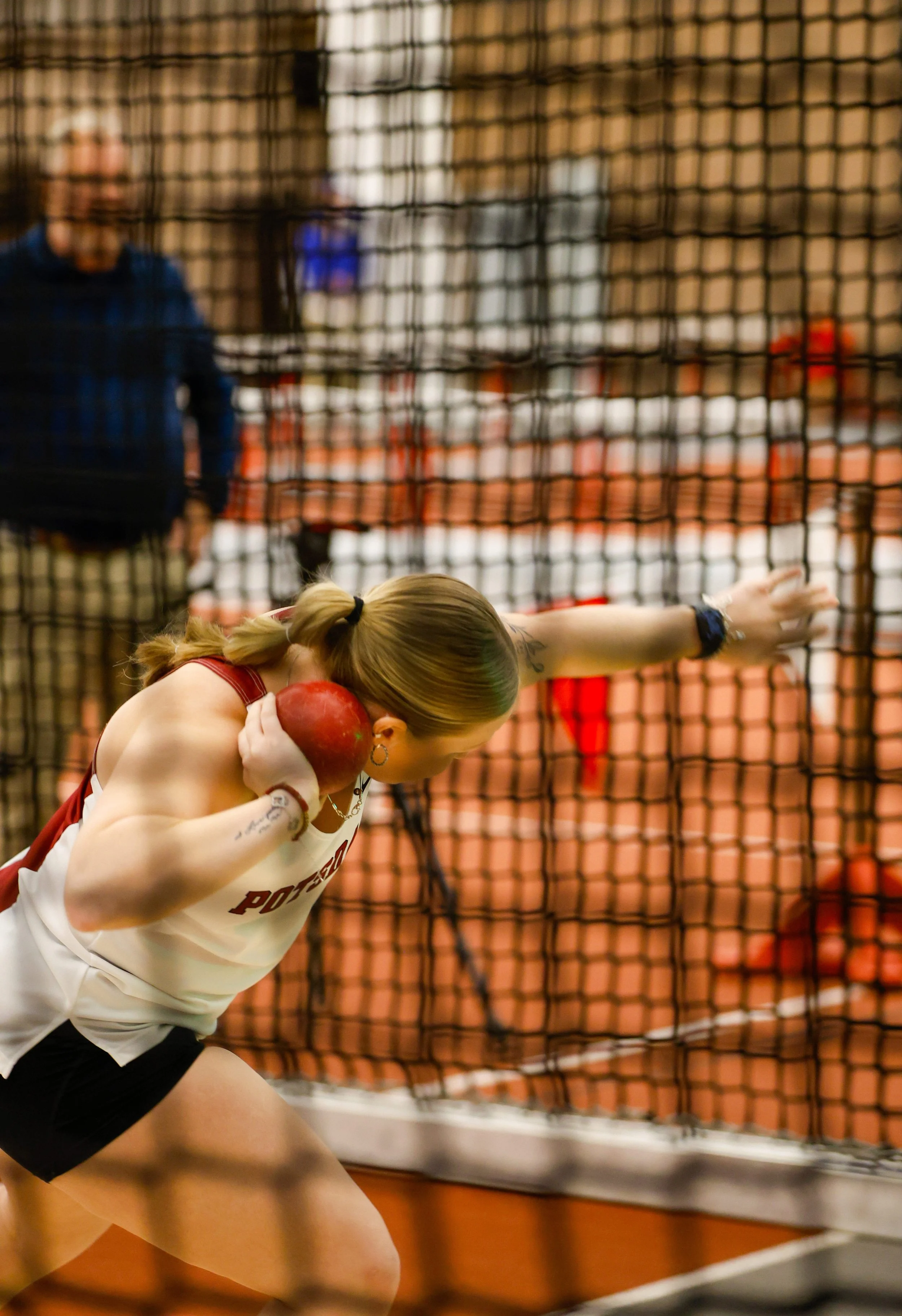 A female athlete in a sports uniform, holding a red ball to her shoulder, preparing to throw on a court with a black net and blurred spectators or players in the background.