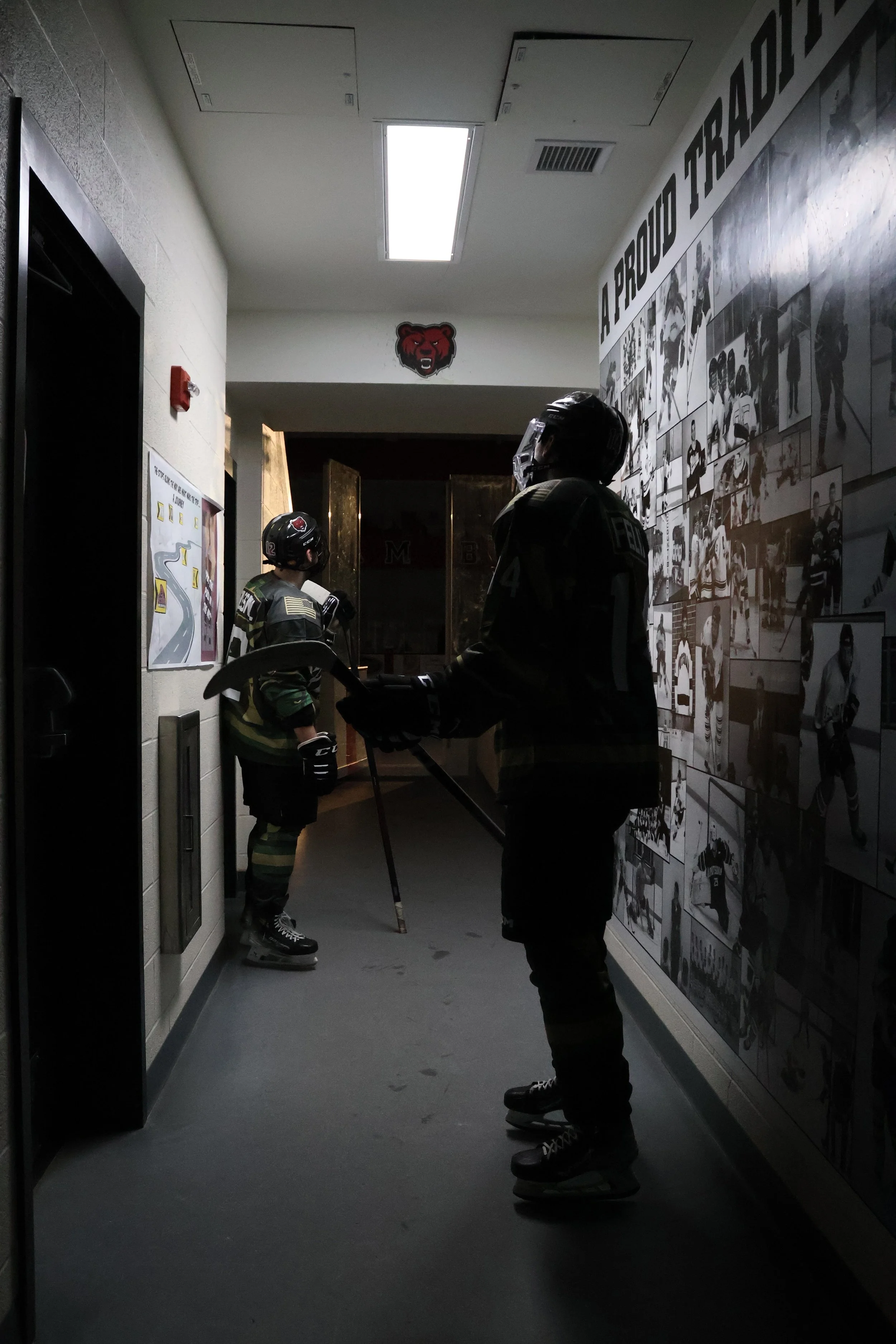 Two people dressed in hockey gear, including skates, helmets, and hockey sticks, standing in a dimly lit hallway with a wall decorated with black-and-white sports photos and a Boston Bruins logo.