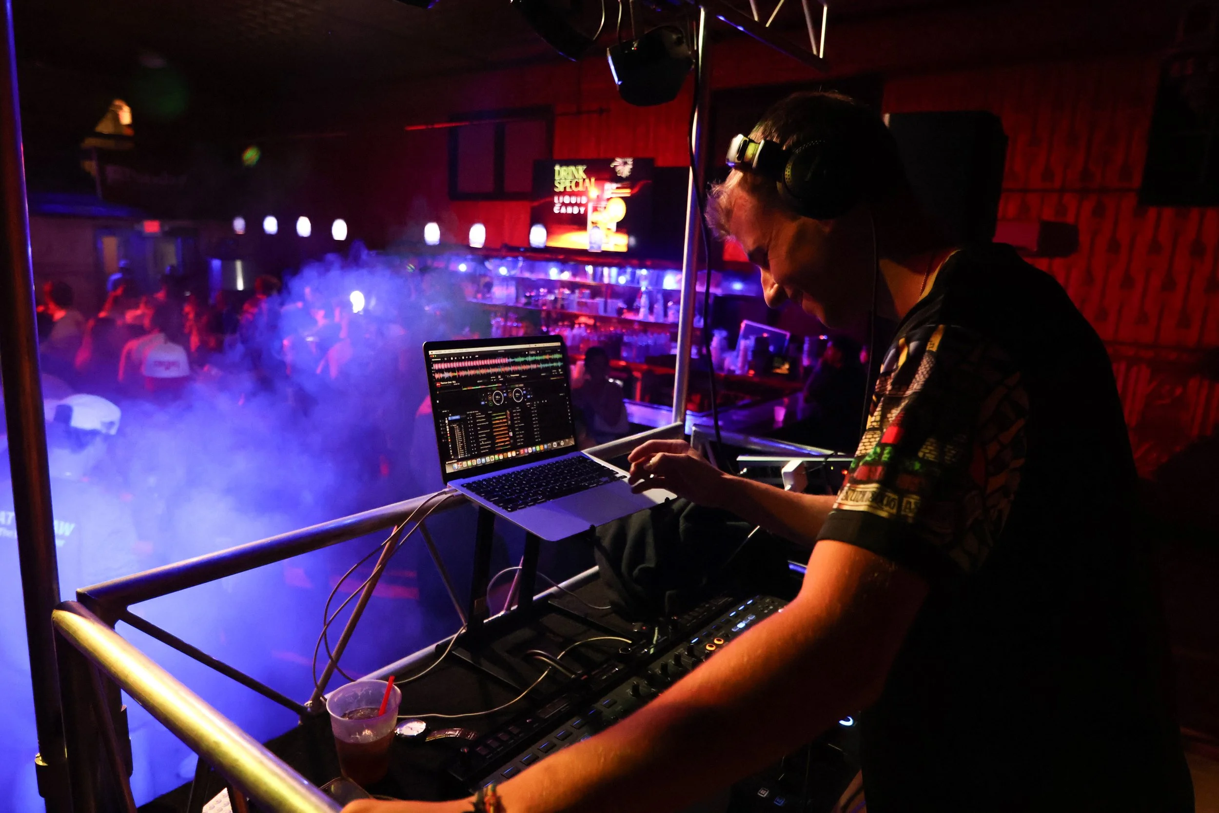 A DJ wearing headphones and a patterned shirt is performing at a nightclub, adjusting controls on DJ equipment with a laptop displaying DJ software. The background is filled with colorful lights, smoke, and a crowd of people dancing in a dimly lit ba