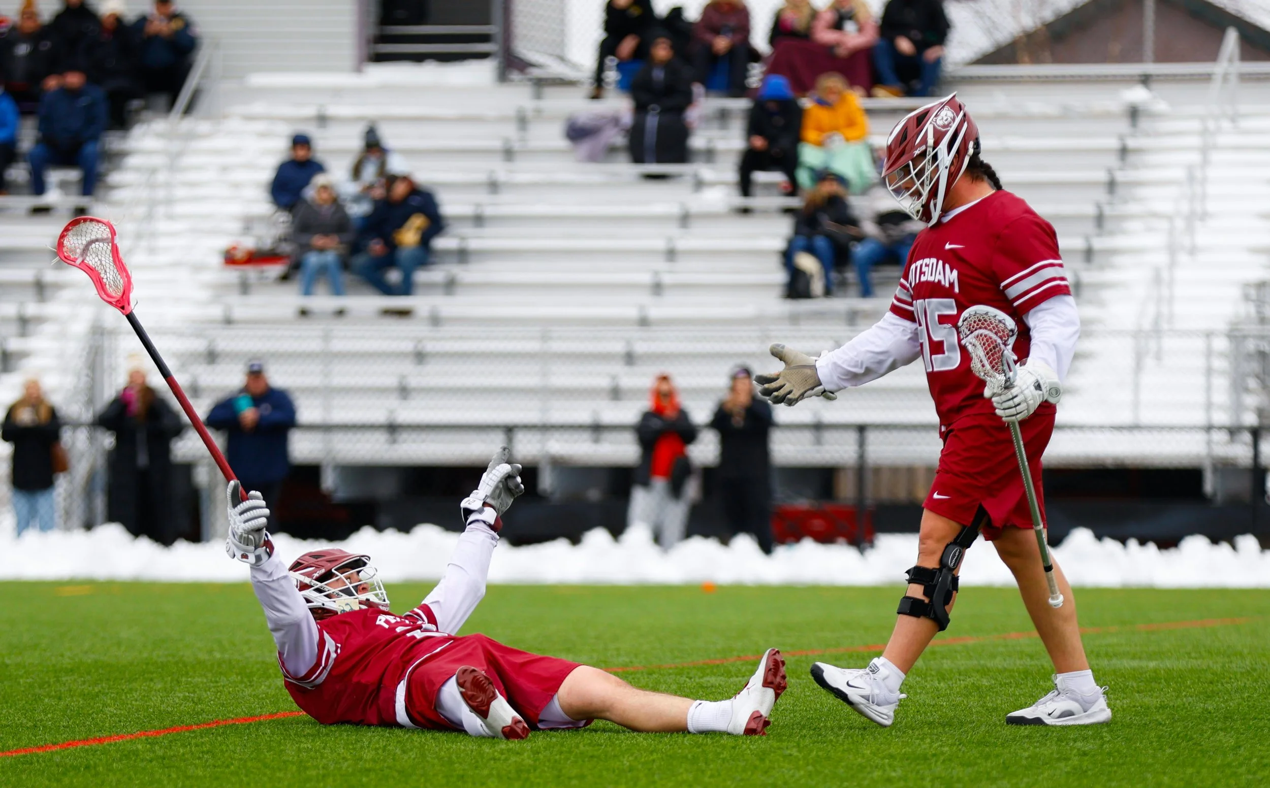 Lacrosse players in red uniforms celebrating on a green field, with empty bleachers and spectators in the background.