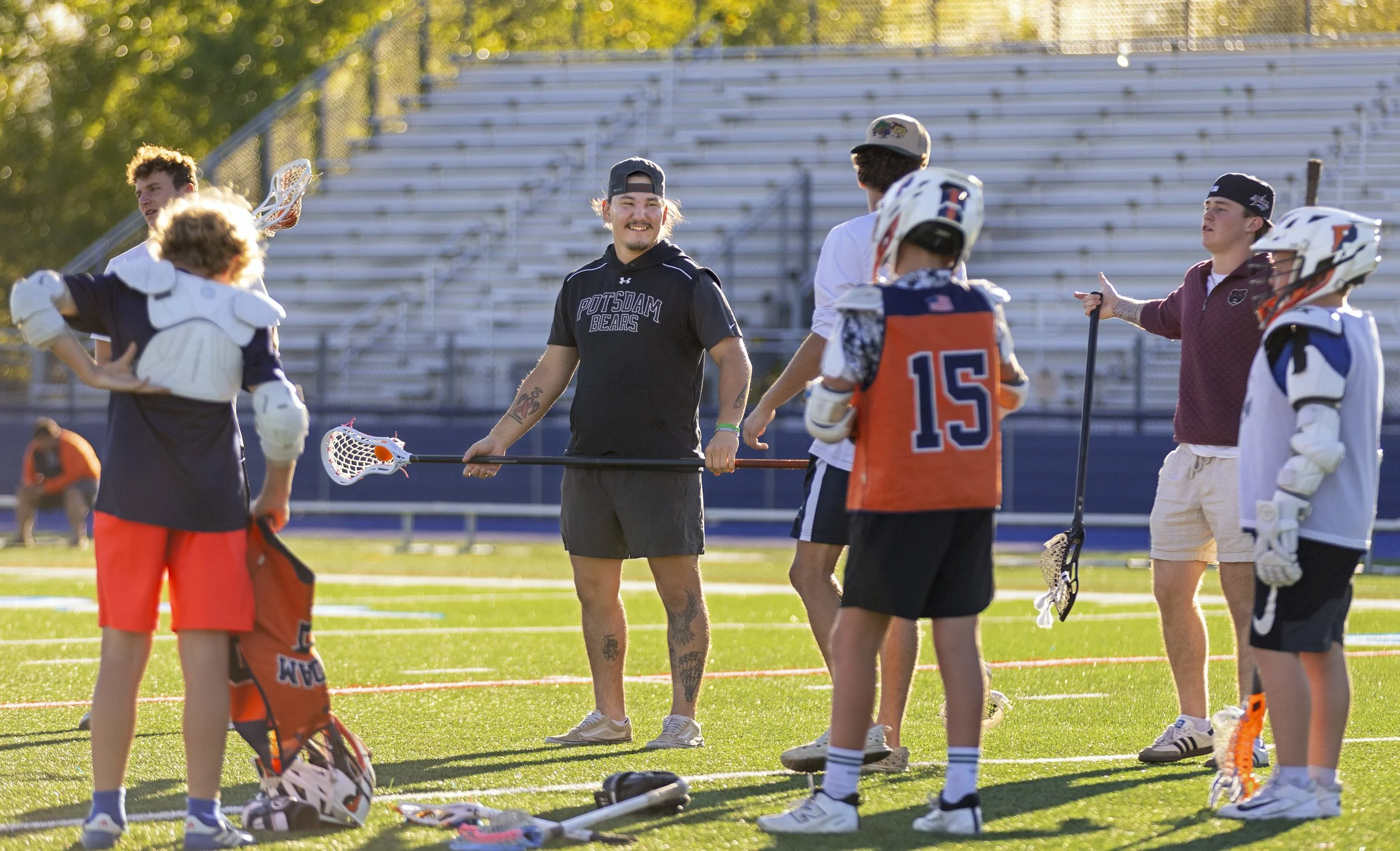 Lacrosse players and coaches gathering on a field, preparing for practice or a game, with a crowded stadium in the background.