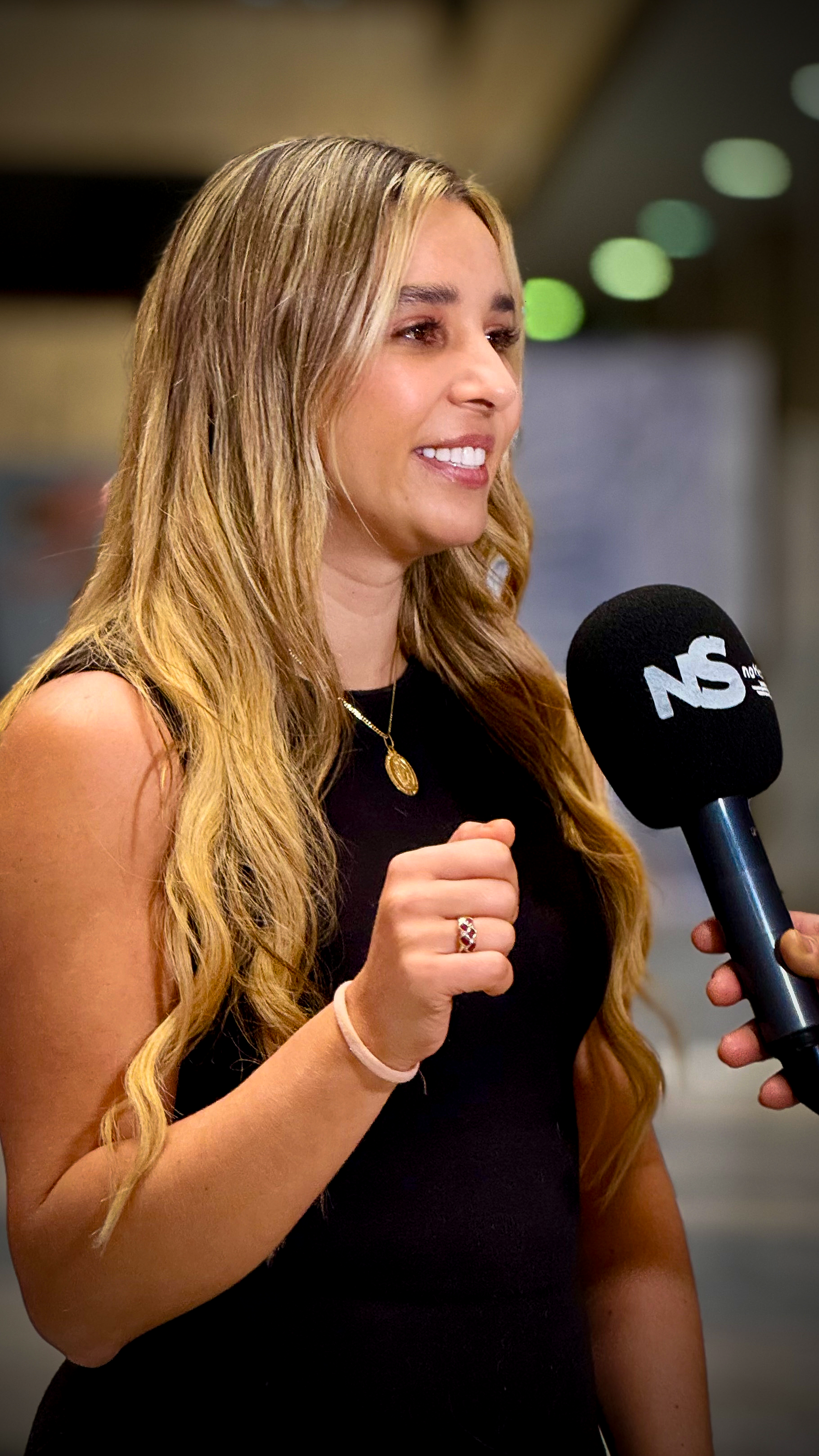A woman with long blonde hair smiling during an interview with a microphone labeled 'NS' in an indoor setting.