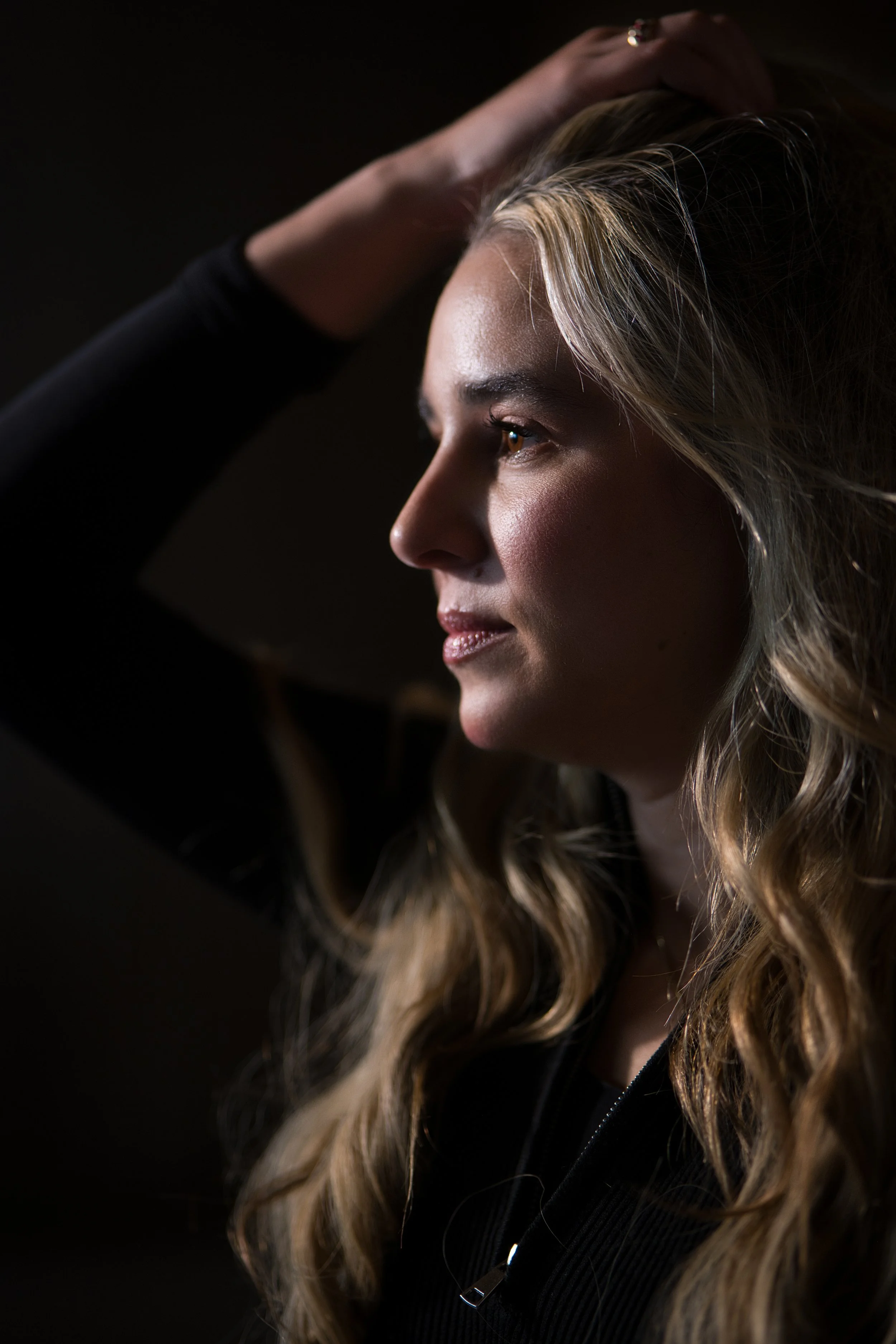 A woman with wavy blonde hair and light makeup is captured in profile, touching her head with her hand, against a dark background.