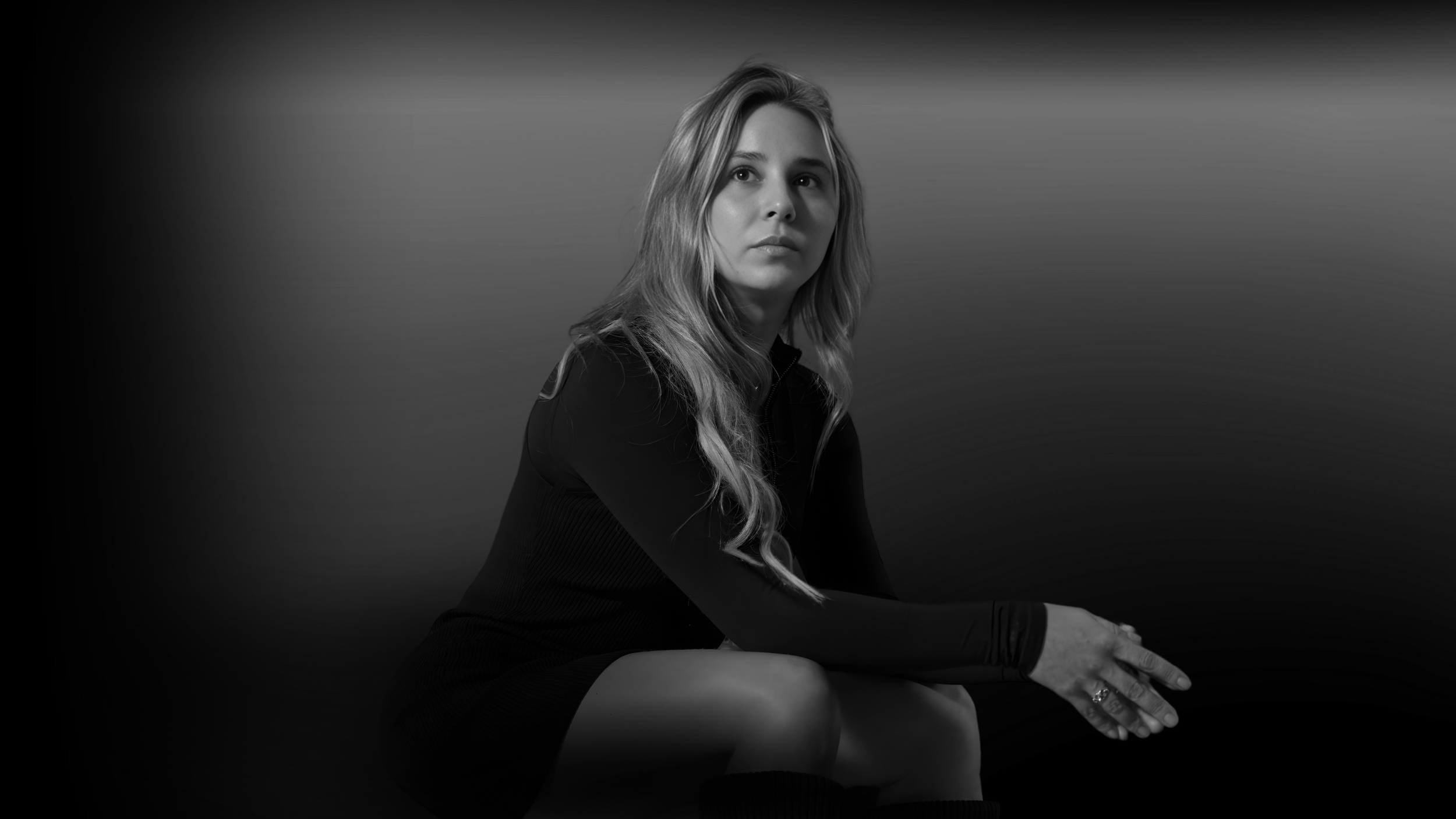 A young woman with long wavy hair sitting down, looking off into the distance, in a black and white photo with a gradient background.