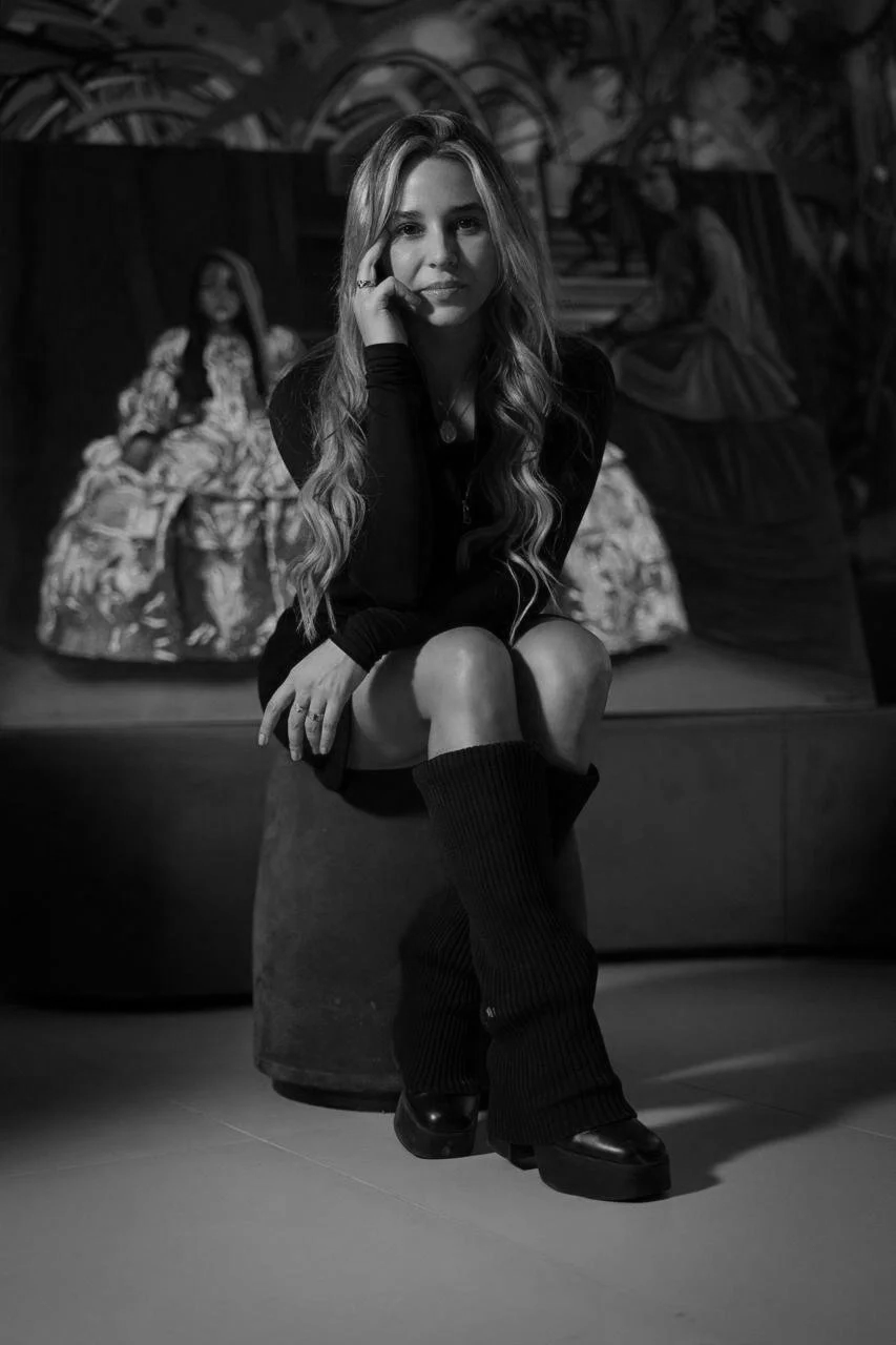 A young woman with long wavy hair sits on a round platform, wearing a black dress or coat and large knit leg warmers with platform shoes. She rests her chin on her hand and looks at the camera. In the background, there are two women in decorative dresses, one sitting and the other sitting on a surface, in a setting with mural or painted design.