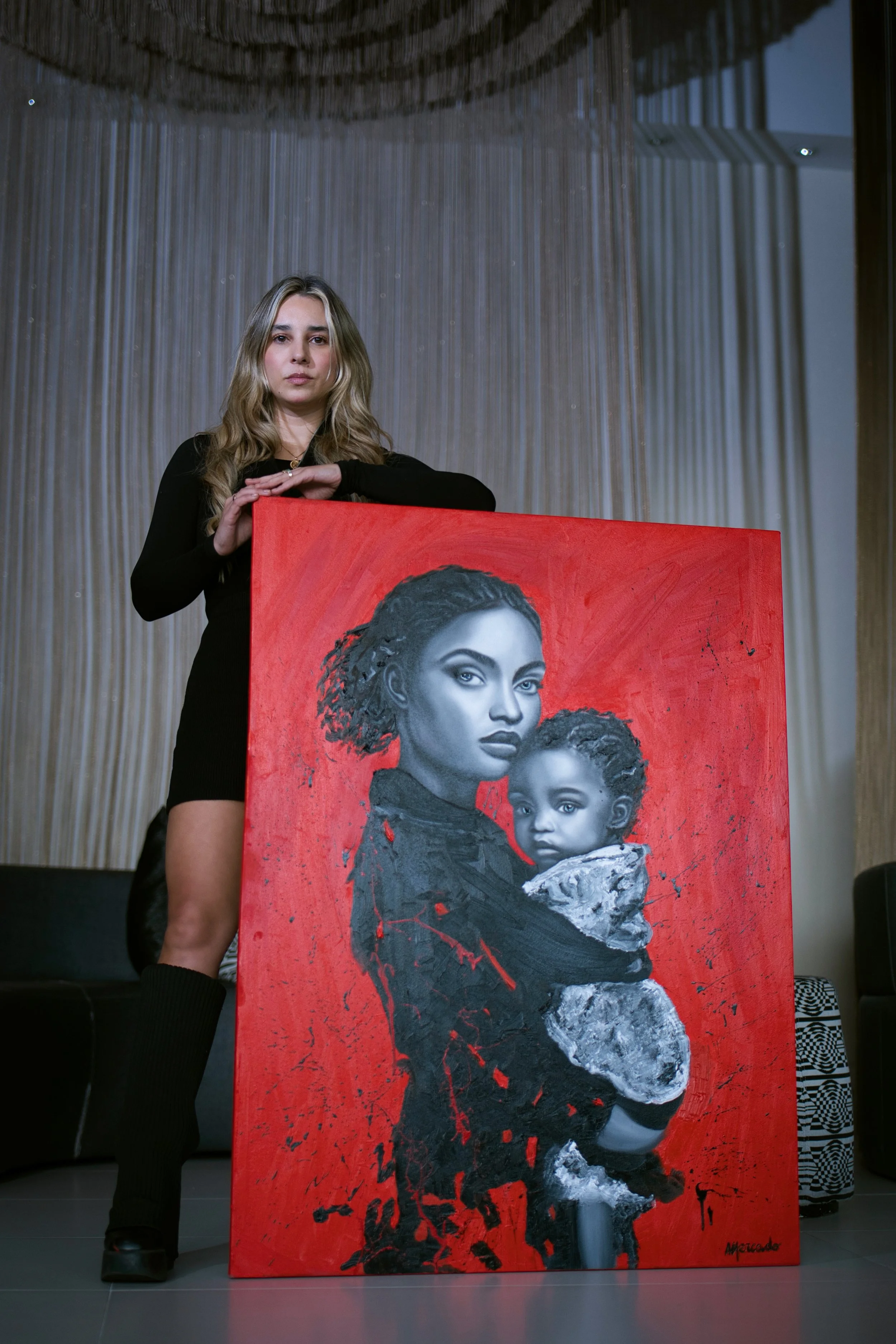 A woman with long blond hair in black clothing stands behind a large painting of a woman and a child, both with dark hair and brown skin, on a red background.