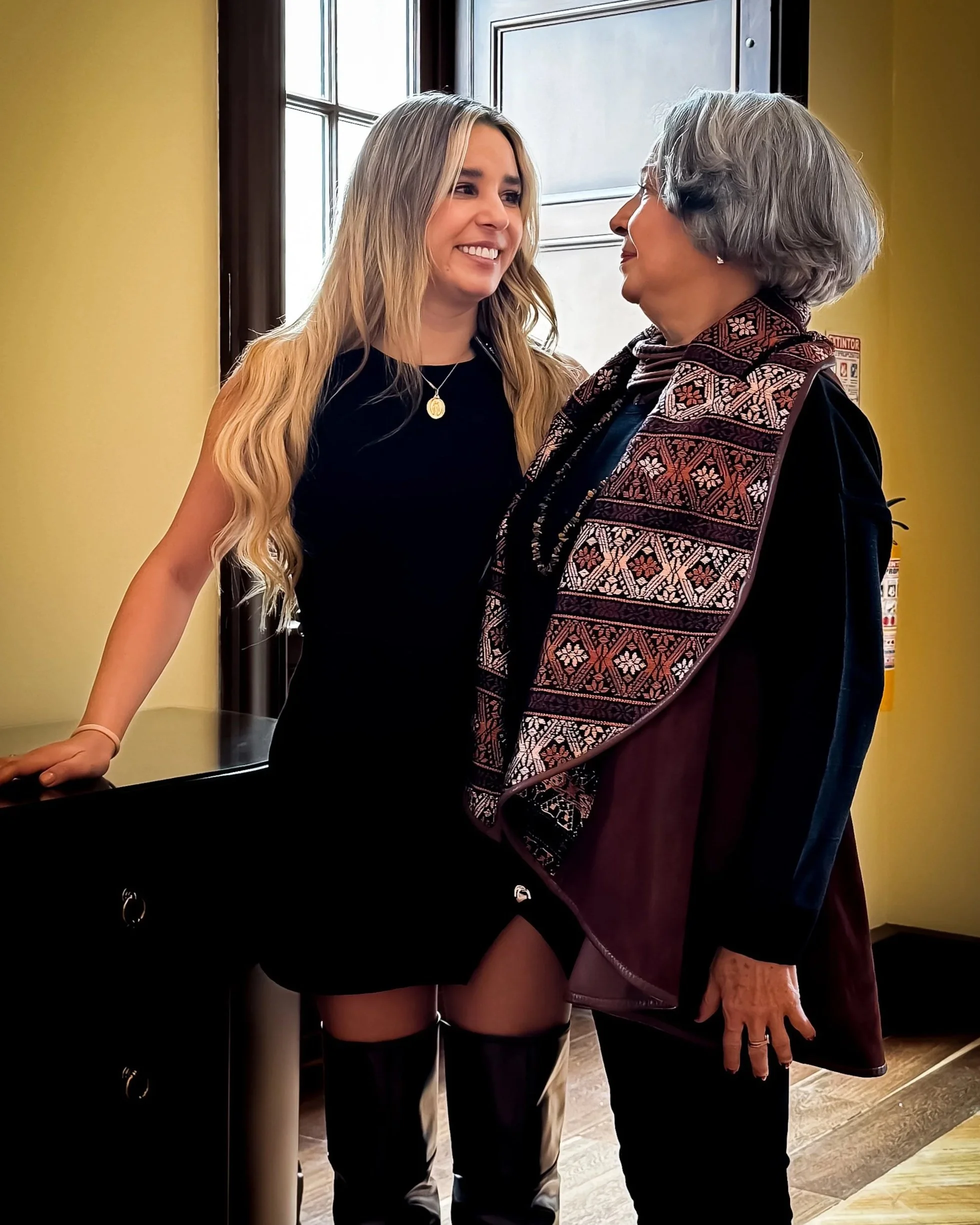 Two women, one younger with long blond hair in a black dress and a gold necklace, and one older with short gray hair in a patterned vest, standing close and smiling at each other in a room with yellow walls and a window.