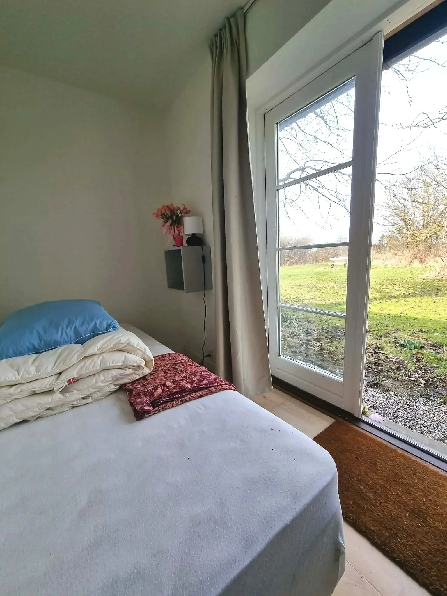 A bedroom with a bed covered in a white sheet, a folded blanket, and a pillow with a blue pillowcase. There is a small wall shelf with pink flowers and a lamp next to a large window showing an outdoor scene of grass and trees.