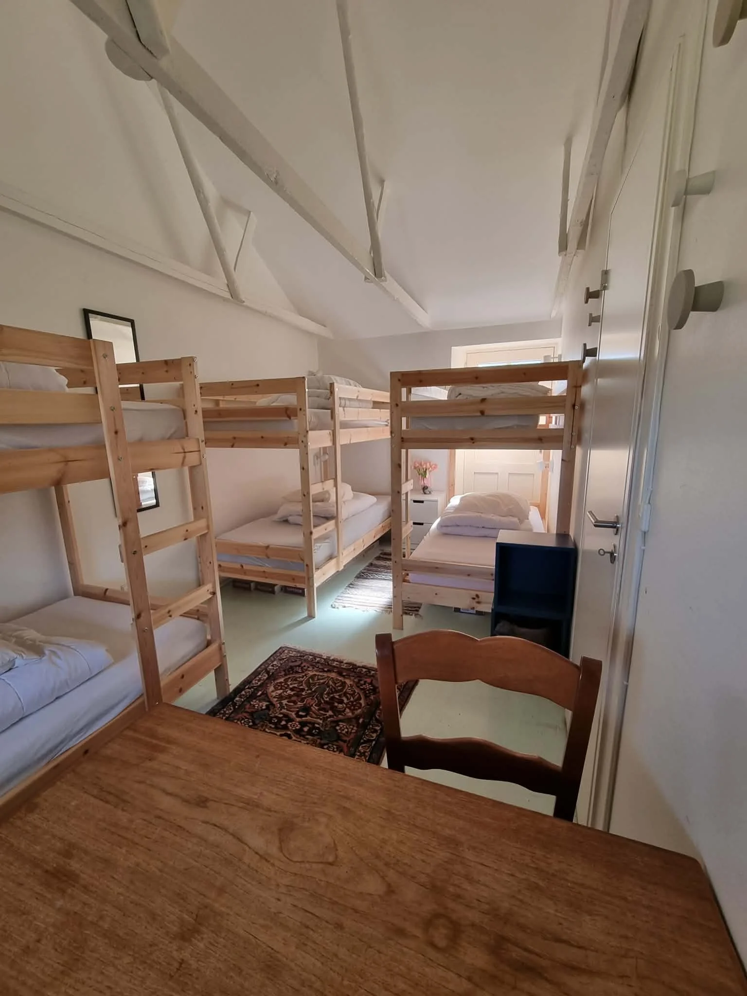 Bedroom with five bunk beds made of light wood, a wooden table and a chair, and a small blue shelf. A patterned rug is on the floor near the beds, and a vase with flowers is on a nightstand near the window.