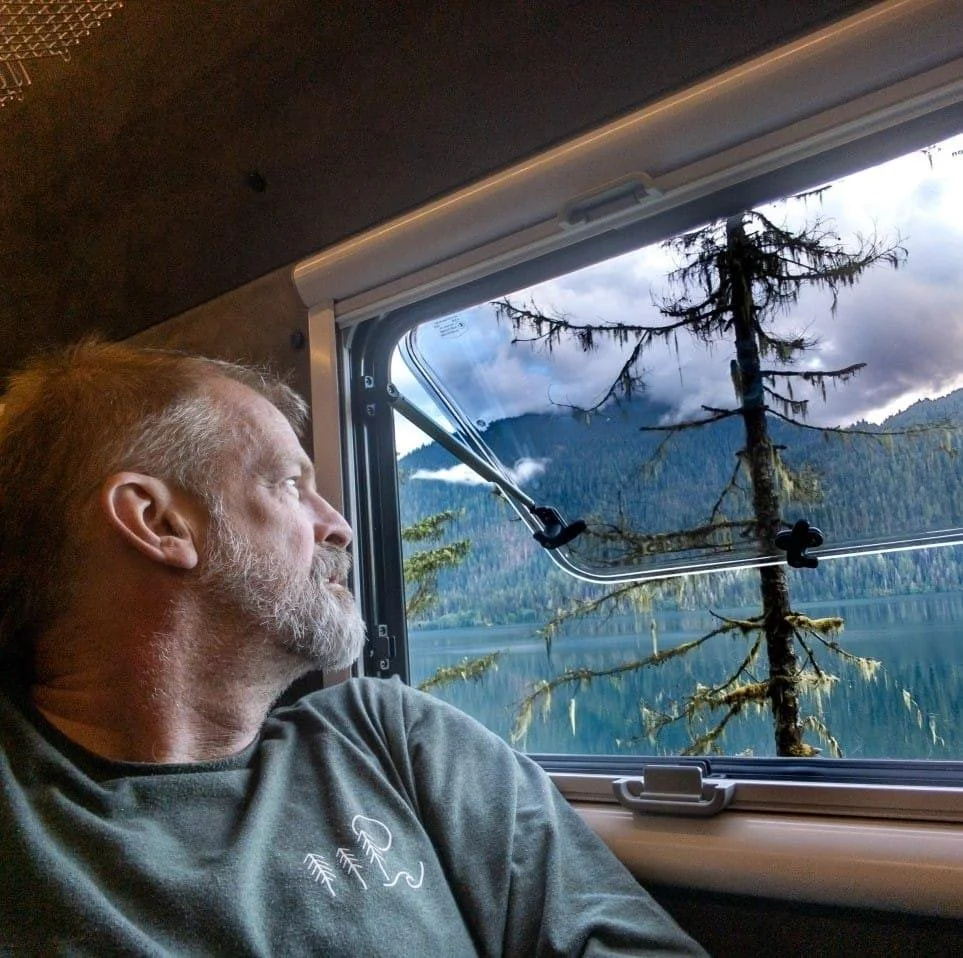 Steve, the photographer, with gray hair and a beard sitting inside his Kimbo Camper, looking out of the window at a landscape with a mountain, a lake, and a tall, moss-covered tree.