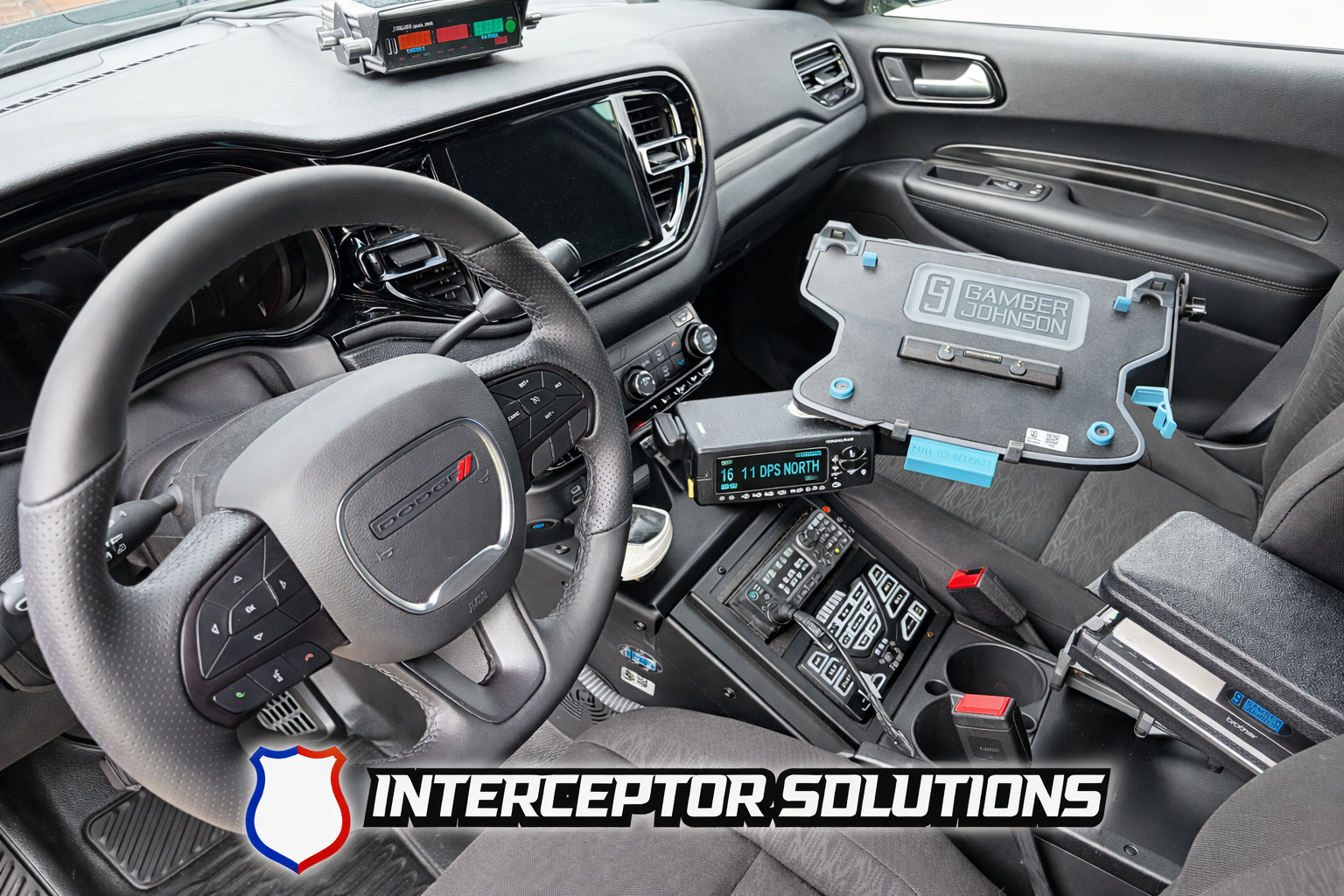 The interior of a police or emergency vehicle with a Dodge steering wheel, multiple communication devices, a mounted device displaying GPS coordinates and direction, a laptop mount with a laptop, and an Interceptor Solutions logo at the bottom.