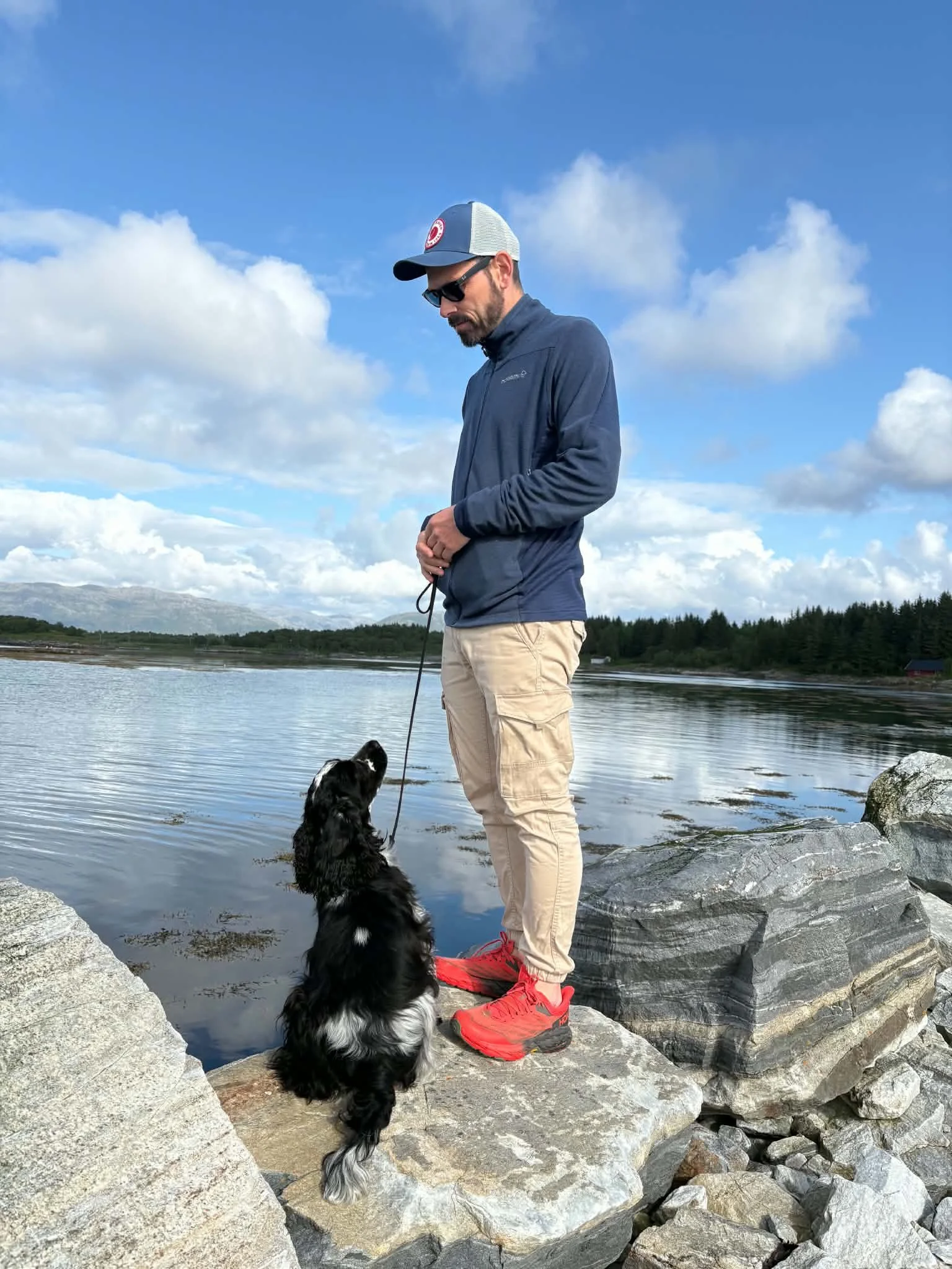 Mann og cocker spaniel står på en stein ved vannet med fjellene i bakgrunnen.