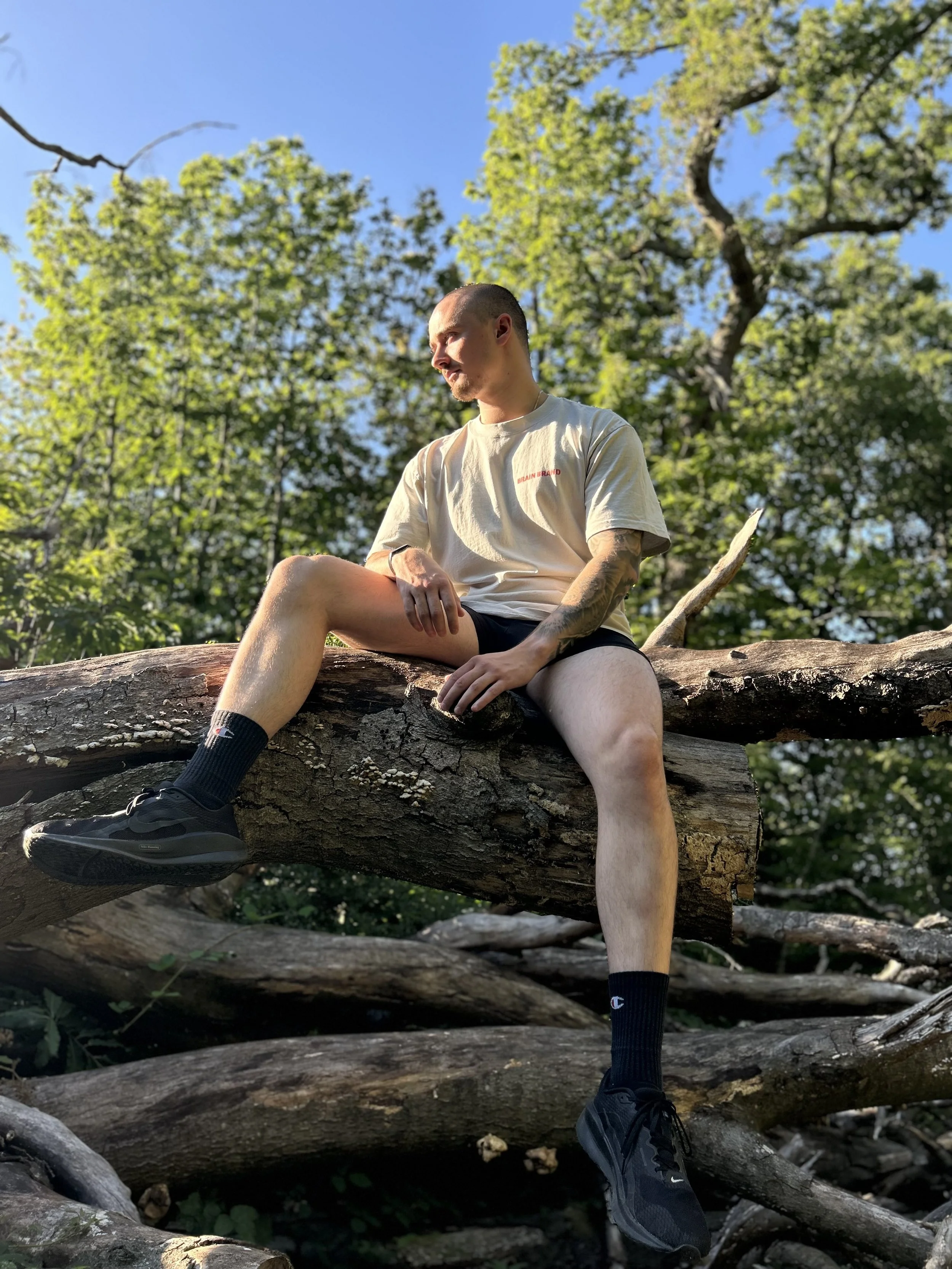 Young man with a bald head and tattoos sitting on a fallen tree trunk in a wooded area with green leaves and blue sky, wearing a beige t-shirt, black shorts, black sneakers, and black socks, appearing relaxed and thoughtful.