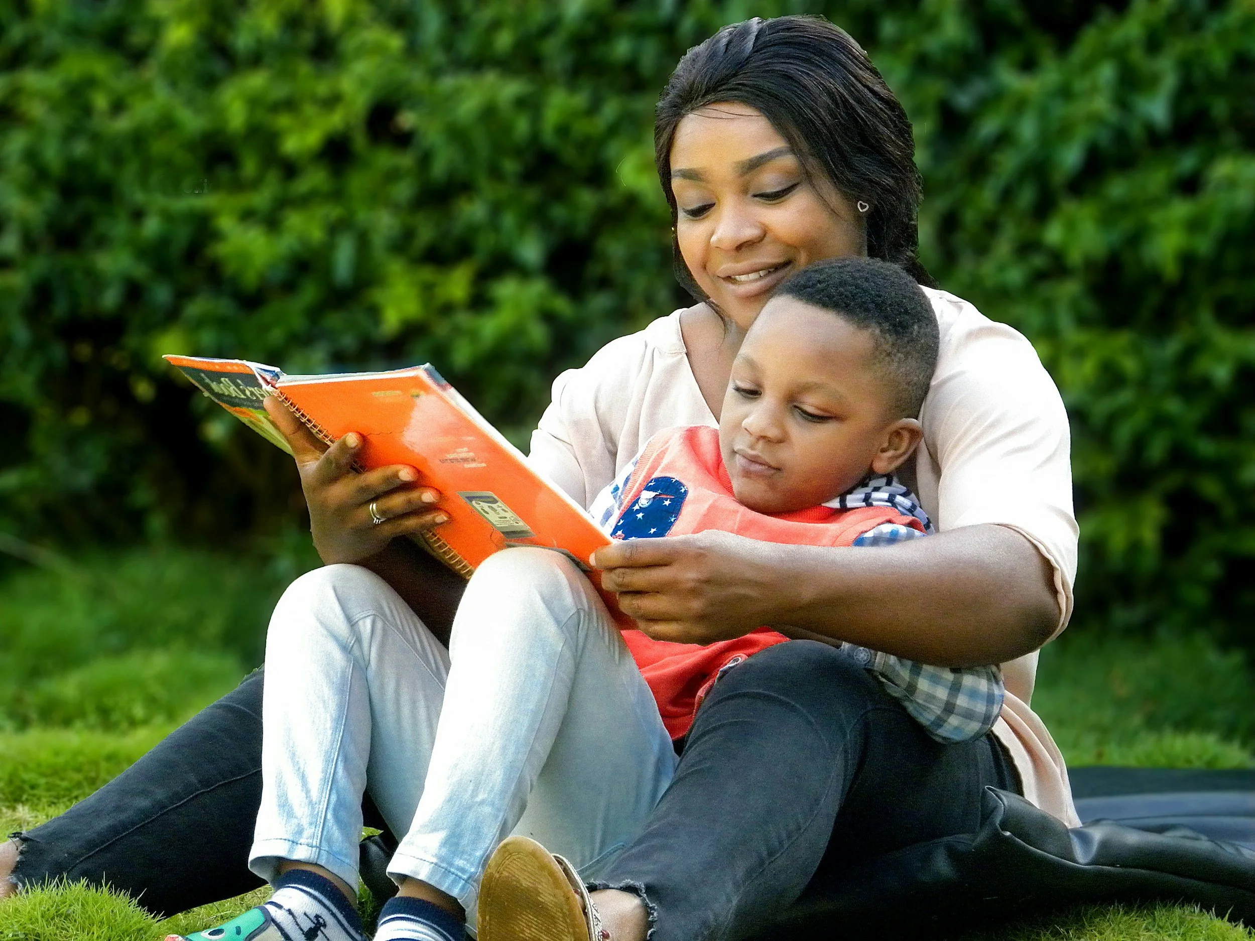 A woman and a young boy sitting on grass outdoors, reading a book together, with green bushes in the background.