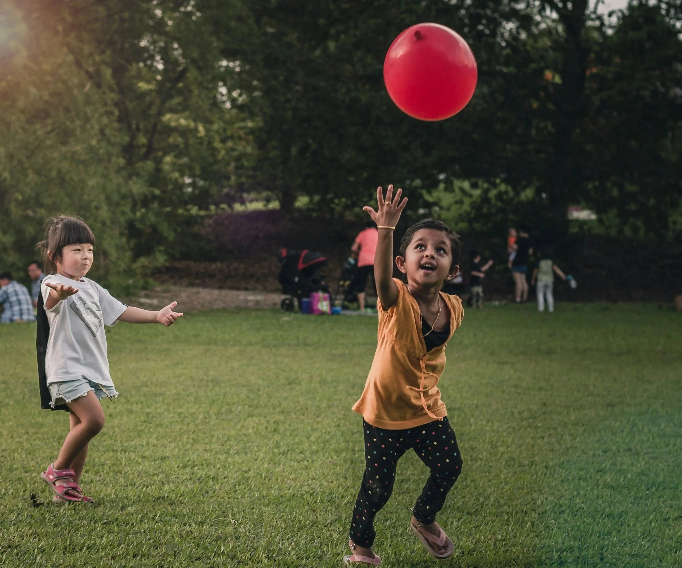 Two children playing with a red ball outdoors on a grassy field during daytime, with other people in the background.
