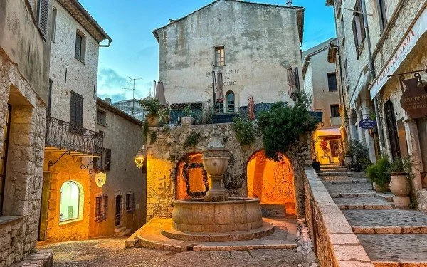 Place ancienne de Saint-Paul-de-Vence avec une fontaine centrale, des maisons en pierre et une lumière chaleureuse en fin de journée.