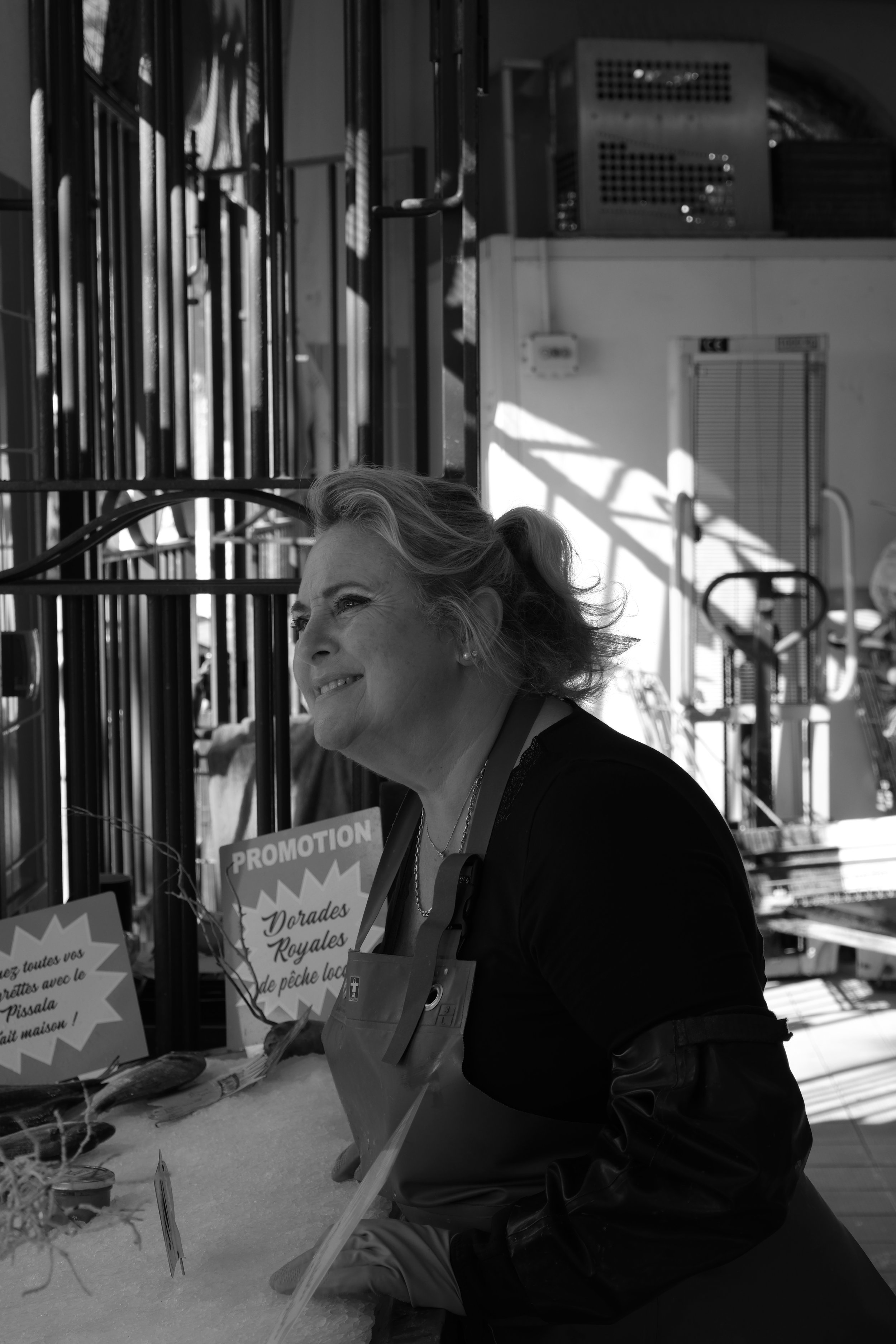 Femme souriante derrière un étal de poissonnerie, dans une scène de marché en noir et blanc.