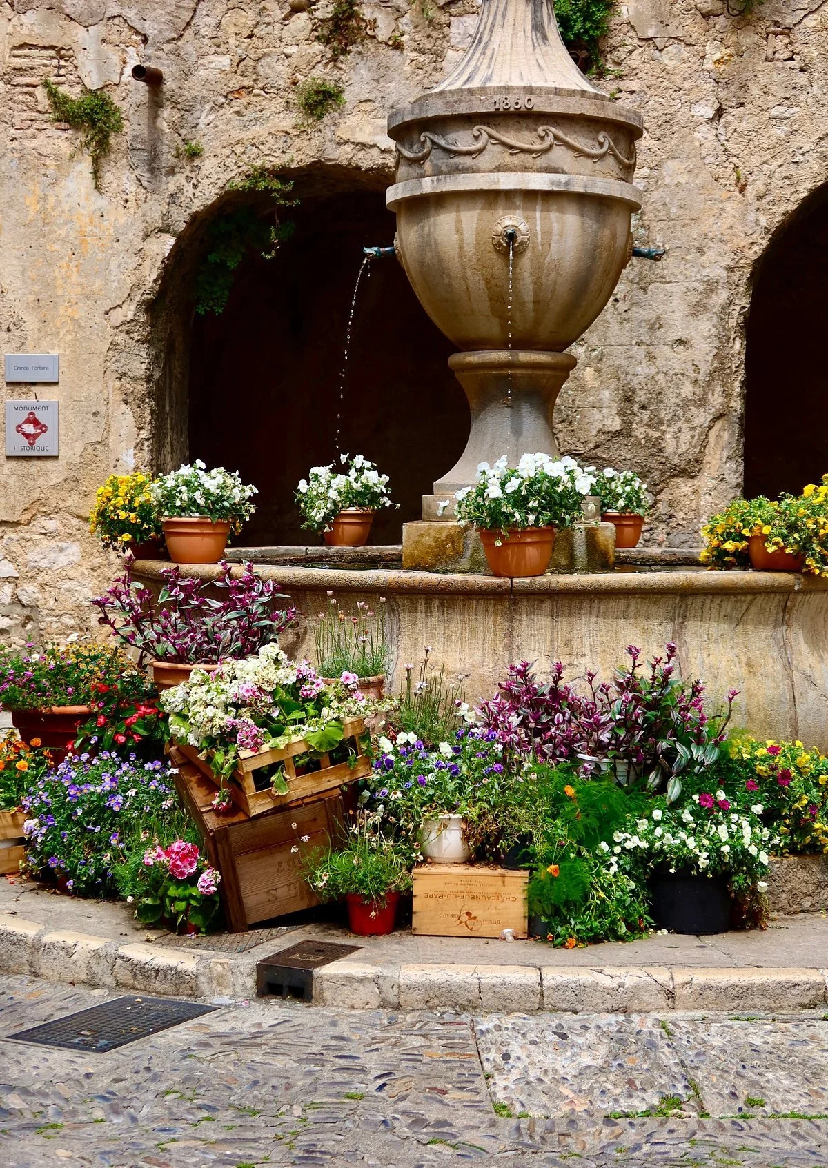 Grande fontaine en pierre entourée de fleurs colorées dans une rue ancienne de Saint-Paul-de-Vence.