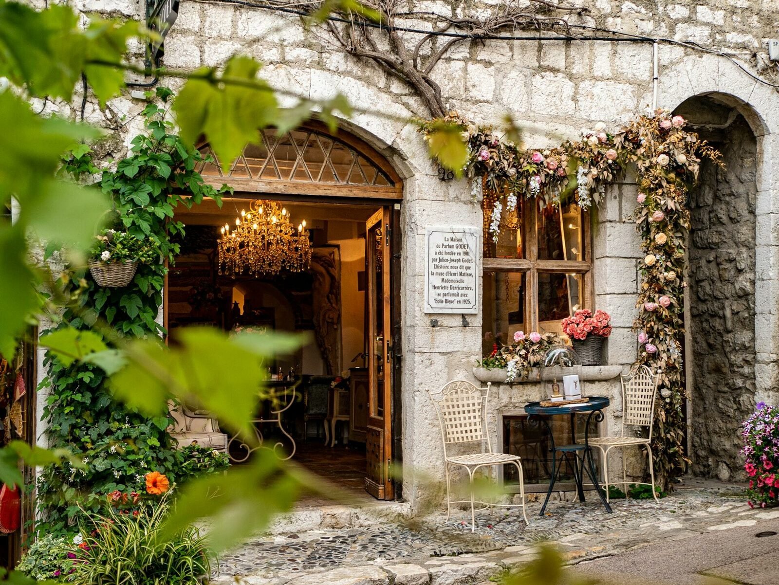 Façade en pierre fleurie avec boutique ouverte, lustre ancien et terrasse décorée dans le village de Saint-Paul-de-Vence.